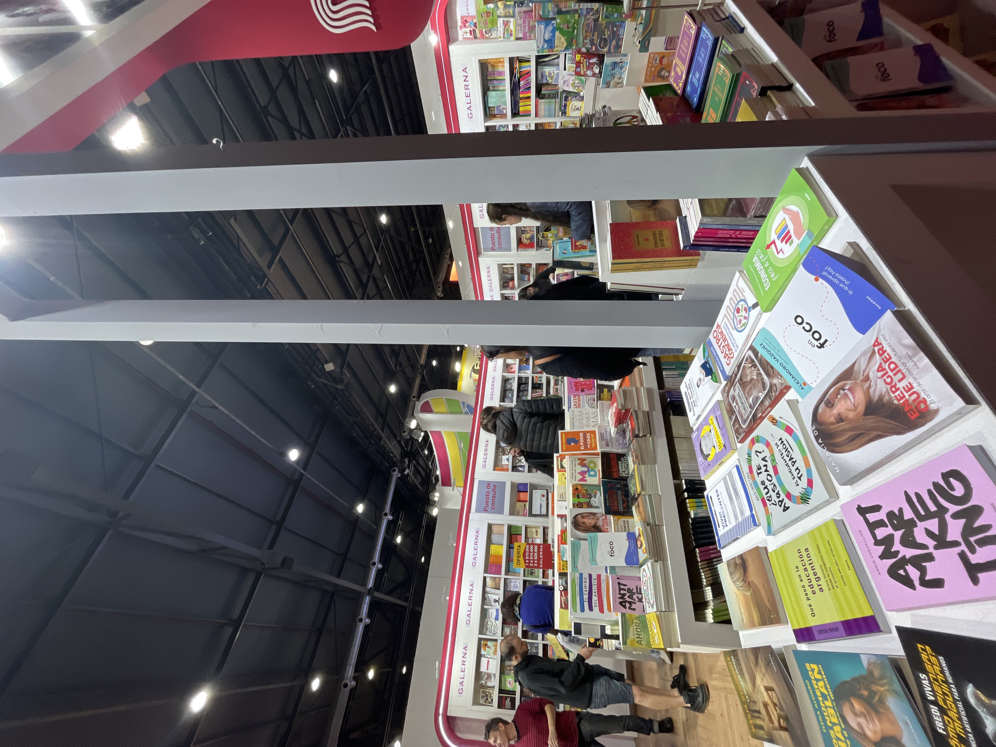 This image shows the "Galerna" stand at the Buenos Aires International Book Fair, located in Buenos Aires, Argentina. There are several bookshelves filled with books. People are browsing the books, reading, and interacting with the exhibition. The environment is well-lit.