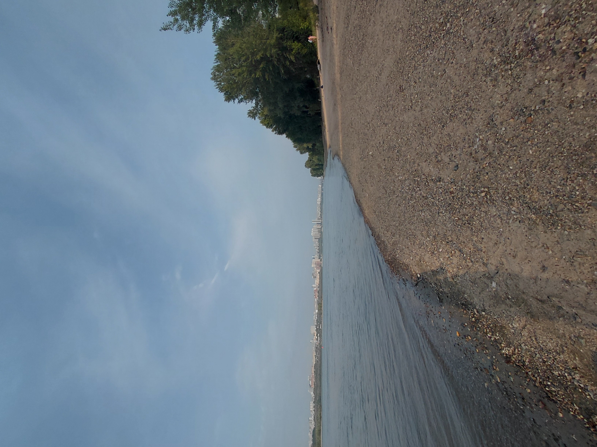 The image depicts a wide river with a city skyline in the distance, likely Novosibirsk, Russia, seen across the water. The foreground is a pebble and sand beach with sparse vegetation. A line of trees borders the beach further down. Two individuals are walking on the beach near the trees. The sky is partly cloudy and the water appears calm.