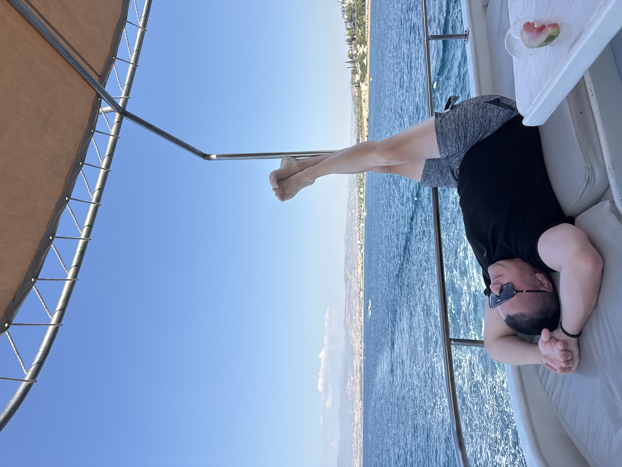 A man is lying on a boat with his legs up in the air. He is wearing sunglasses and a black t-shirt. The boat is on the sea, with a city and mountains in the background. The city appears to be Neo Chorio, Cyprus. The sky is clear and blue.