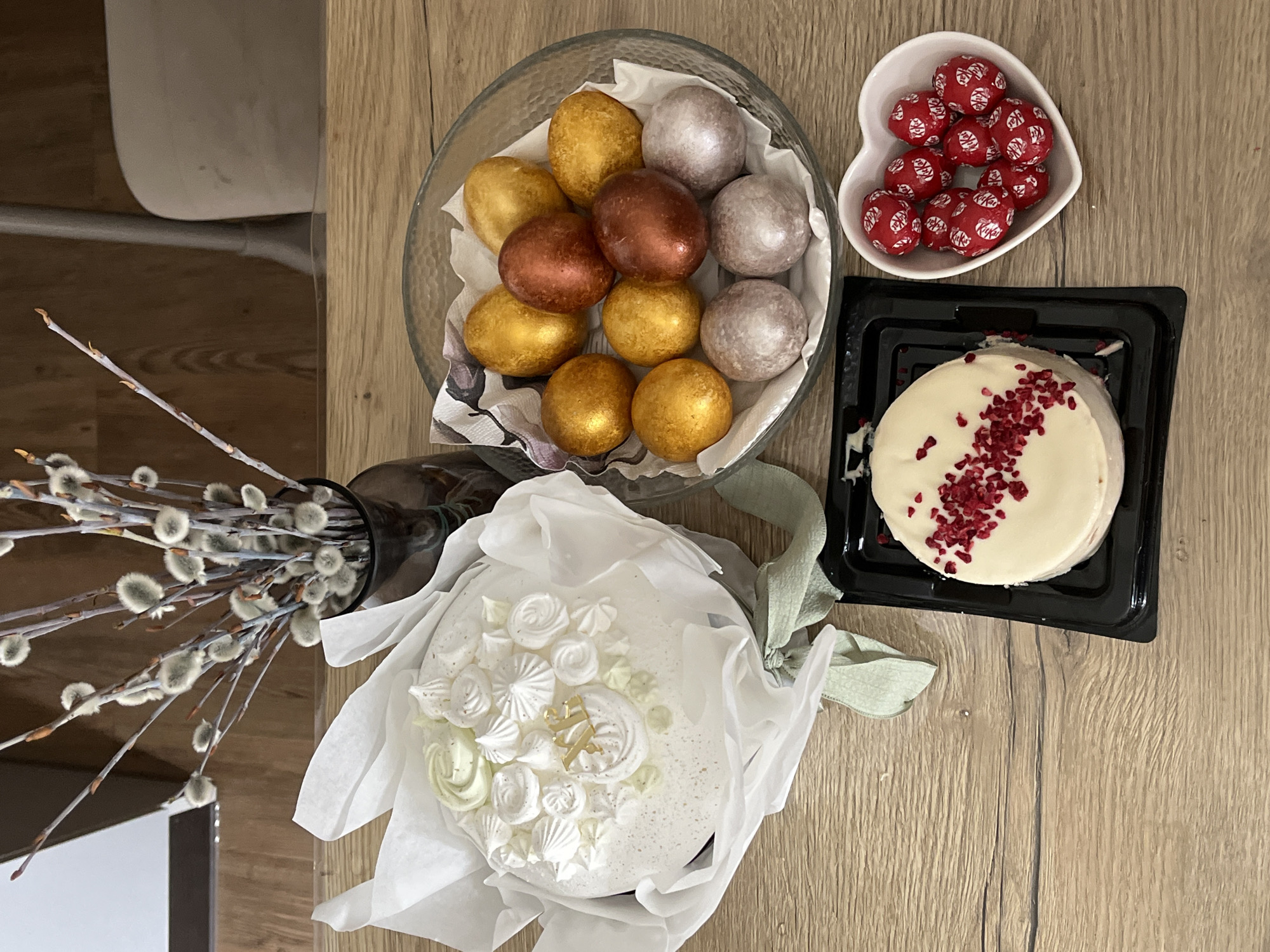 A top-down shot shows a wooden table displaying Easter treats and decorations. A clear glass bowl is filled with gold, bronze, and silver colored Easter eggs. A heart-shaped white bowl contains red Kit Kat balls. A small round cake with white icing and red berry sprinkles sits in a square black container. Another cake, decorated with white icing, meringue swirls, and gold sprinkles, is wrapped in white paper and tied with a light green ribbon. A dark brown vase holds willow branches with fluffy catkins. The location is unspecified, and there is no confirmed association with Krasnoyarsk, Russia. No people are visible.