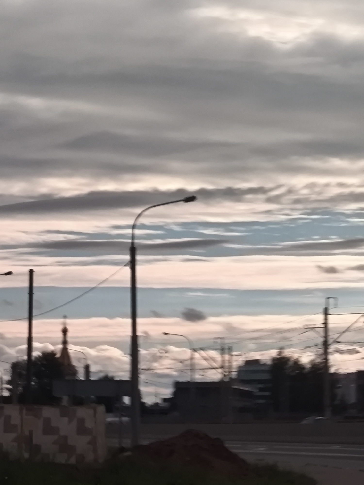 The image depicts an outdoor scene under a cloudy sky. The sky is filled with horizontal bands of clouds, varying in color from dark grey to light pink and blue, suggesting it might be near dawn or dusk. Silhouetted against the sky are several streetlights on tall poles, with power lines crisscrossing between them. In the middle ground, a church steeple with a golden dome is visible, surrounded by trees and buildings. The foreground shows a street with a pile of dirt on the right side, and a patterned wall on the left. The overall impression is one of a city landscape at an indeterminate time of day, possibly during a transitional period of light. There is no visible text in the image.