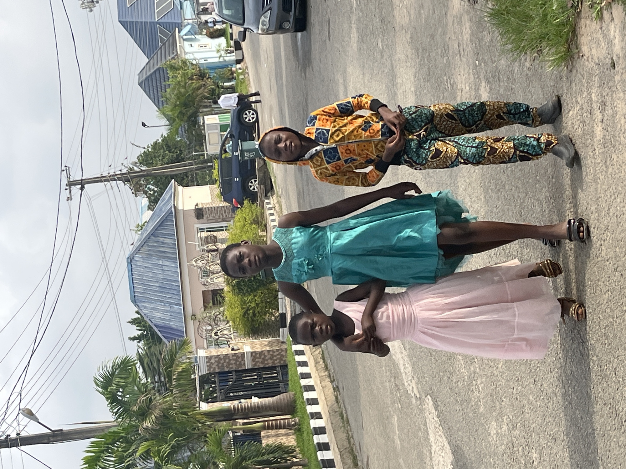 Three young girls stand on a paved street. The girl on top wears a yellow and blue patterned jacket and pants. The middle girl wears a light blue dress, and the bottom girl wears a light pink dress. The background includes buildings with blue roofs, a black SUV, trees (including palm trees), and utility poles with wires. The location is likely in a residential area of Abuja, Nigeria.
