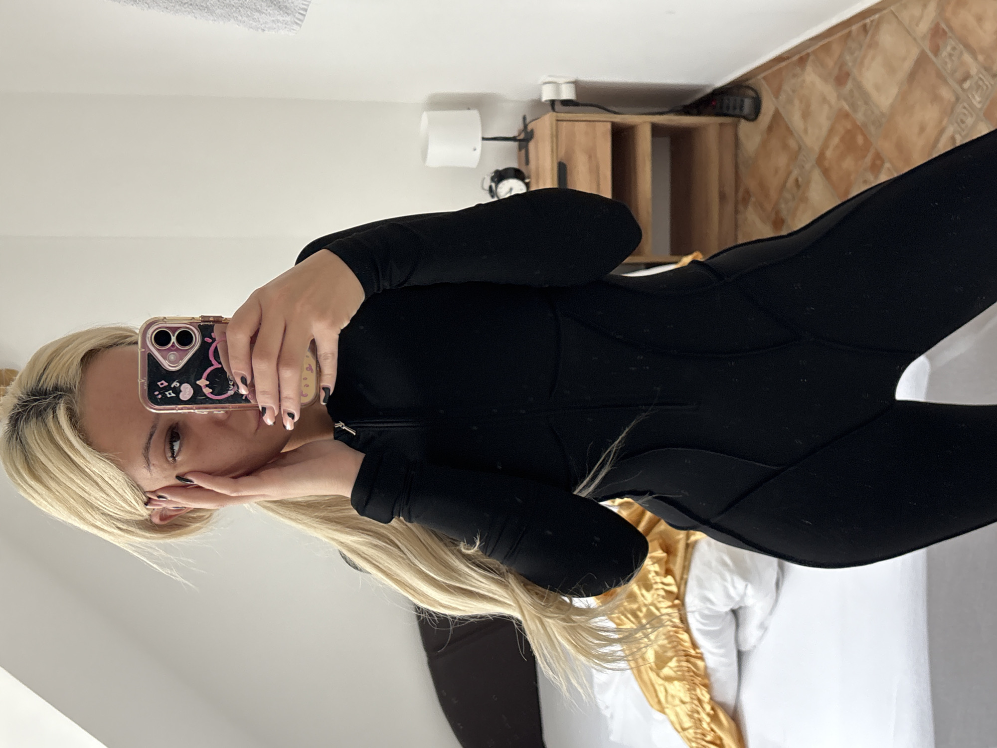 A young woman is captured in a mirror selfie indoors, appearing to be in a bedroom or hotel room. She has long, straight blonde hair and fair skin, with her light-colored eyes looking directly into the camera. She is wearing a form-fitting, long-sleeved black jumpsuit or bodysuit, which features subtle paneling details. Her left hand holds a smartphone, while her right hand is raised, partially obscuring her mouth and chin. Her nails are manicured, with some painted black and others a lighter shade with black tips.
The smartphone is encased in a translucent pink or purple cover adorned with black cartoon designs, possibly a cat. The dual camera lenses of the phone are clearly visible.
The background of the room features plain white walls. On the right side of the image, a light wooden bedside table or cabinet with a dark metal frame holds a white, cylindrical lamp, a black analog alarm clock with white hands, and a dark-colored remote control. The floor is covered with light brown and beige square tiles arranged in a diagonal pattern. A portion of a bed with white bedding and a golden, ruffled throw blanket is visible in the lower right corner.
The activity taking place is a self-portrait captured in a mirror. The overall mood is casual and focused on the photograph. No specific time of day or weather is indicated. There is no discernible text visible in the image. The setting, with its tiled floor and wooden furniture, aligns with the context of Sofia, Bulgaria.