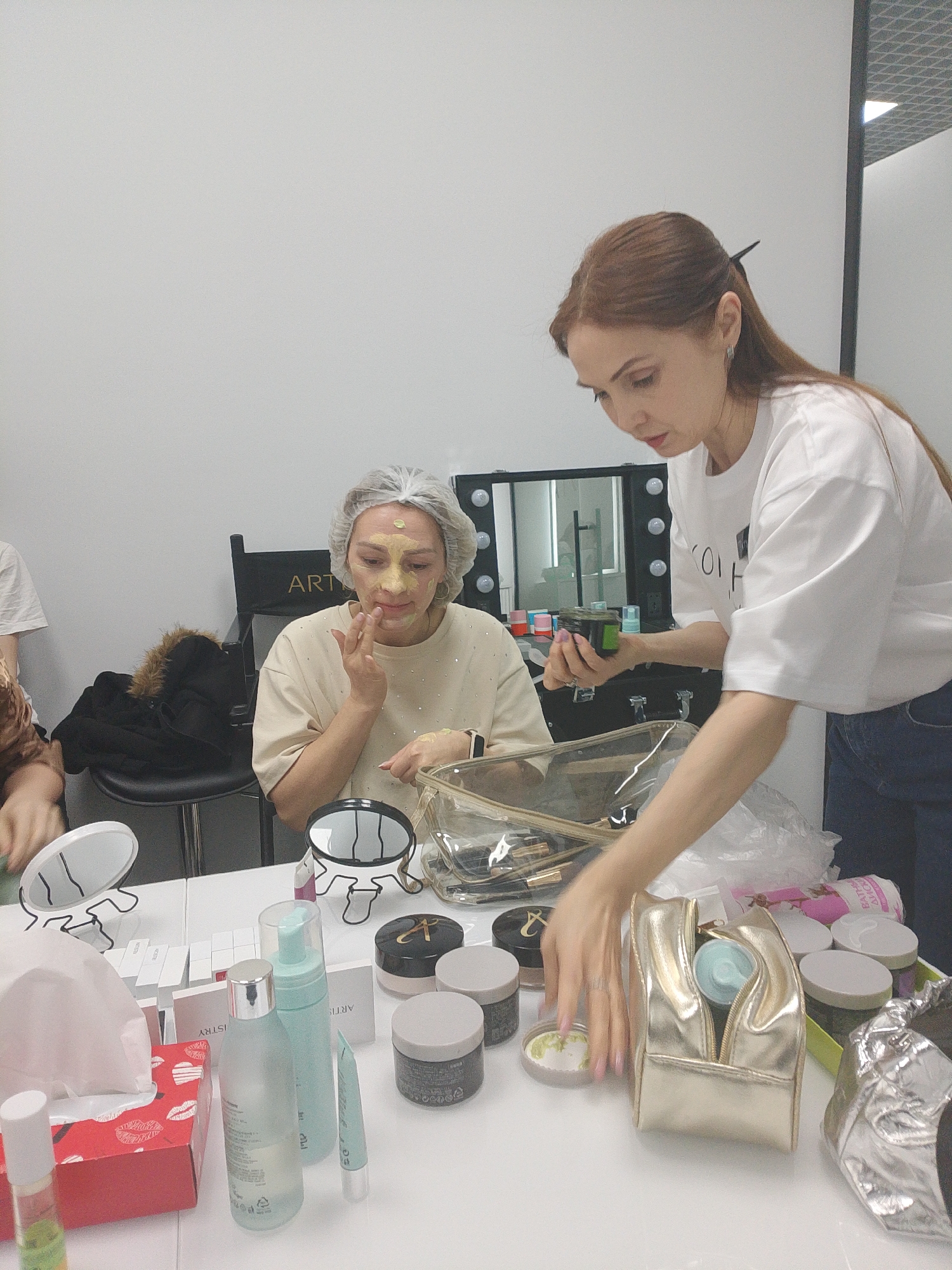 In a brightly lit interior space, two women are the primary subjects of the photograph. One woman is seated at a table, wearing a beige shirt and a white hair covering, with a yellow substance applied to her face. She is touching her face with her right hand. The other woman stands next to the table, facing the seated woman, holding a green container in her left hand, and wearing a white shirt and blue jeans. She has reddish-brown hair pulled back with a clip.
The table is cluttered with various beauty products, including jars, bottles, and makeup bags. A tissue box with a red and white floral design is visible. Behind the seated woman, there is a director's chair with the word "ARTIST" written on it, and a mirror with light bulbs surrounding it. A black coat is draped over the chair.
The location may be inferred to be Izhevsk, Russia. The image composition suggests a professional setting, possibly a beauty salon or makeup studio.
