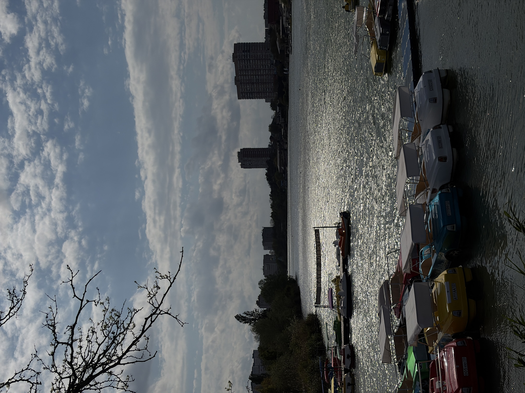 The image depicts a scene on a sunny day at a lake or large pond in Krasnodar, Russia. The water surface is reflecting sunlight, creating a shimmering effect. Along the right edge of the image, there is a dock with a row of colorful paddle boats lined up. Beyond the water, on the opposite bank, are buildings, trees, and other vegetation. In the foreground on the left, there is a bare tree branch. The sky is partially cloudy with a mix of blue sky and white clouds. Tall apartment buildings are visible in the background, confirming a developed urban environment.