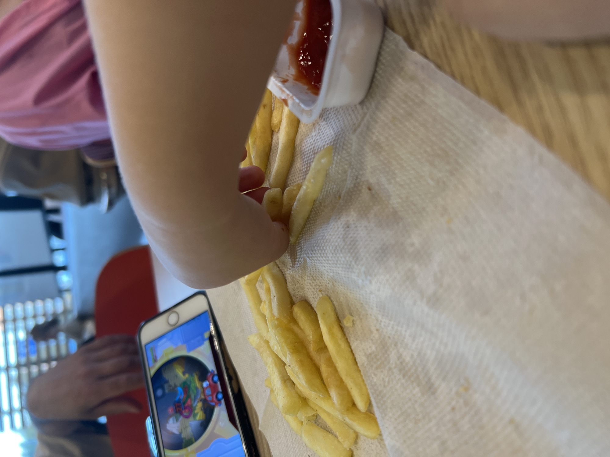 In this close-up, low-angle shot, a person's arm, clad in a pink garment, is reaching towards a serving of French fries. The fries are spread out on a white paper towel, and a small white container of ketchup is tilted over them, with some ketchup dripping onto the fries. A portion of a smartphone is visible in the foreground, displaying a colorful game interface with cartoonish characters and a red vehicle. The background is blurred, showing hints of furniture and a light-colored wall, possibly in a restaurant or home setting. The overall lighting suggests an indoor environment, likely during the day, as natural light seems to be present. The scene evokes a casual dining experience, possibly with a child or a young adult enjoying a meal. There is no visible text in the image.