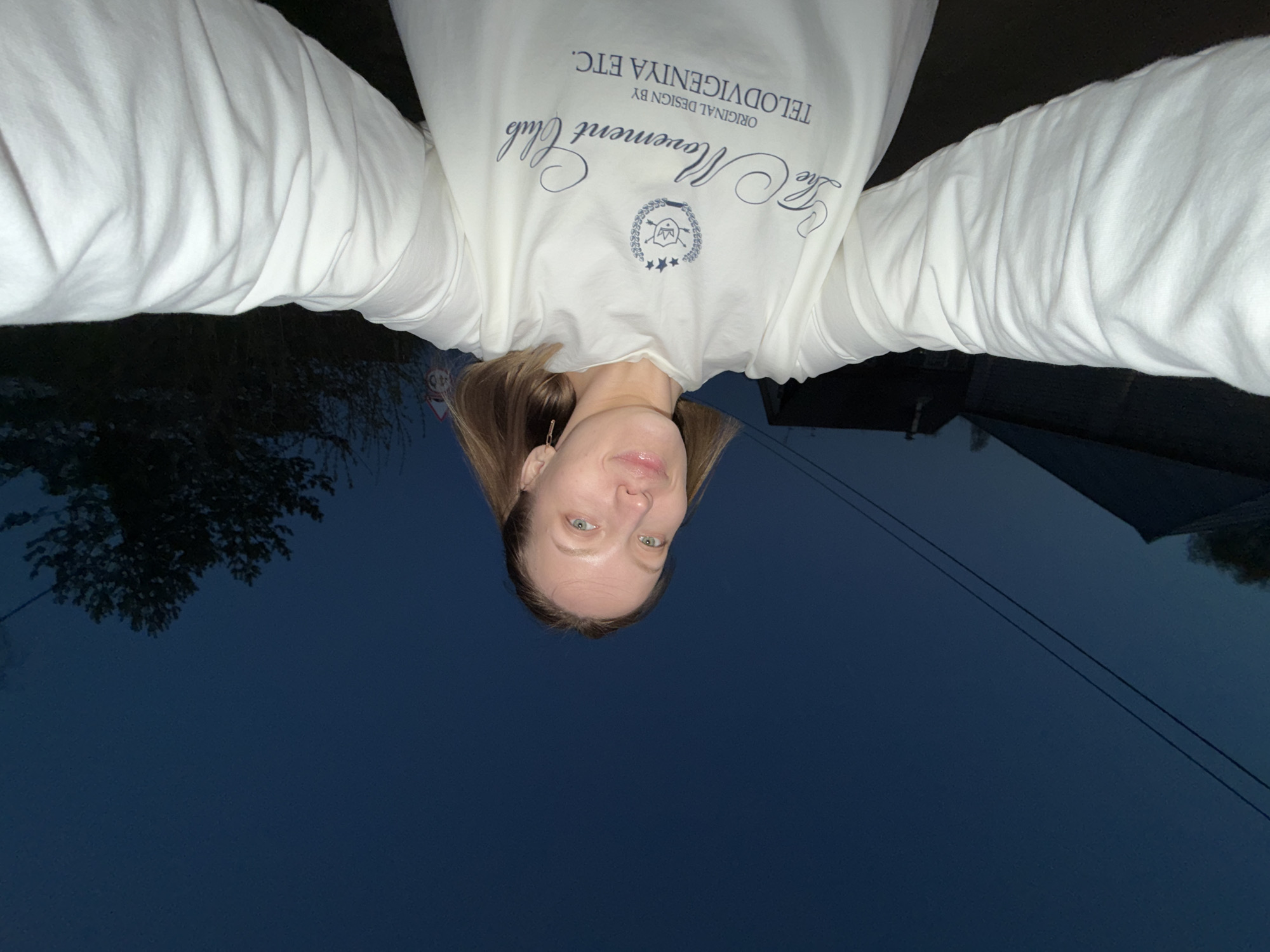 A selfie, possibly taken at dusk or twilight, shows a woman with fair skin and light hair, her face centered and upside-down in the frame. She is wearing a white long-sleeved shirt with navy blue lettering; the text reads "The Movement Club" and "ORIGINAL DESIGN BY TELODIVIGENIYA ETC.". The background features a mix of dark blue sky, trees, and building silhouettes. The location may be Reshetnikovo, Russia, due to the camera originating from this location.