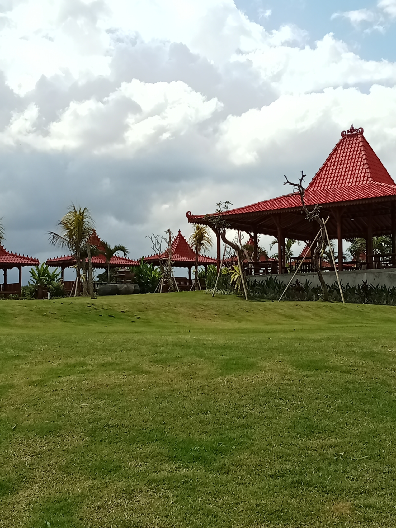 The image depicts an outdoor scene with traditional Indonesian architecture in Selemadeg Timur, Indonesia. In the foreground, a lush green lawn slopes gently upwards. The middle ground features several open-air gazebos with distinct red, multi-tiered roofs, characteristic of Balinese style architecture. These structures appear to be set on a slightly elevated platform, bordered by greenery and some barren trees. Palm trees are visible interspersed among the gazebos, suggesting a tropical climate. The background is dominated by a dramatic sky filled with a mix of white and grey clouds, indicating potentially overcast or changing weather conditions. There are no people visible in the frame. The scene conveys a sense of tranquility and cultural heritage.
