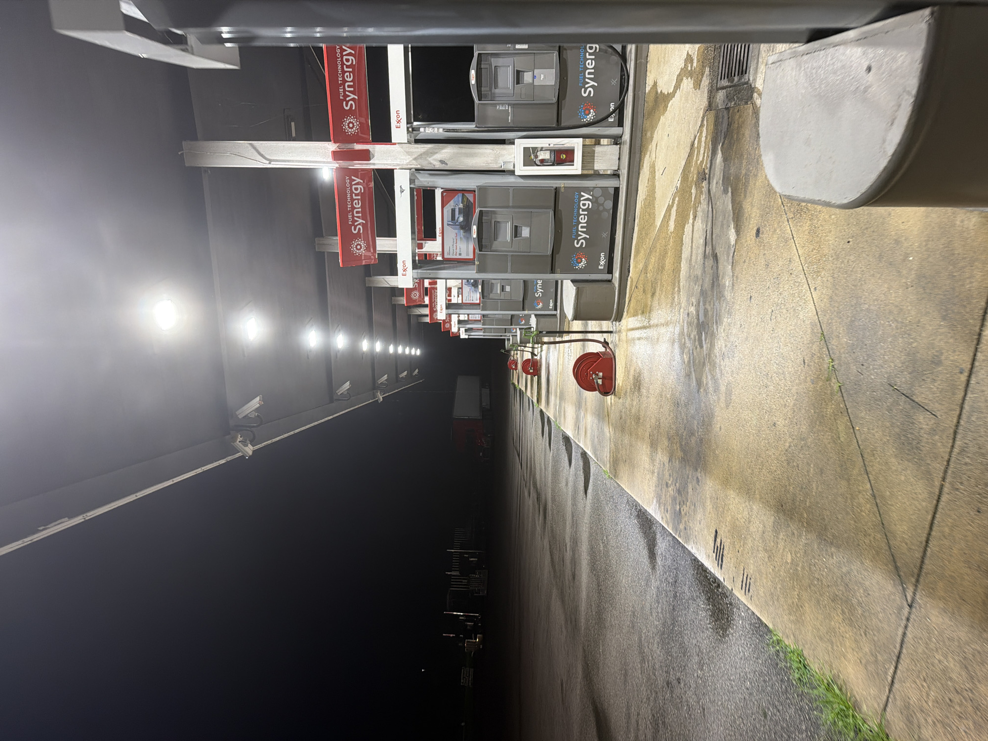 The image depicts an outdoor scene at night, likely a gas station, in Wildwood, United States. A row of illuminated gas pumps is visible under a covered structure with overhead lighting. The ground appears wet, with reflections of the lights. In the background, a semi-trailer truck is parked in the darkness. The overall impression is one of a quiet, possibly deserted, service area.
