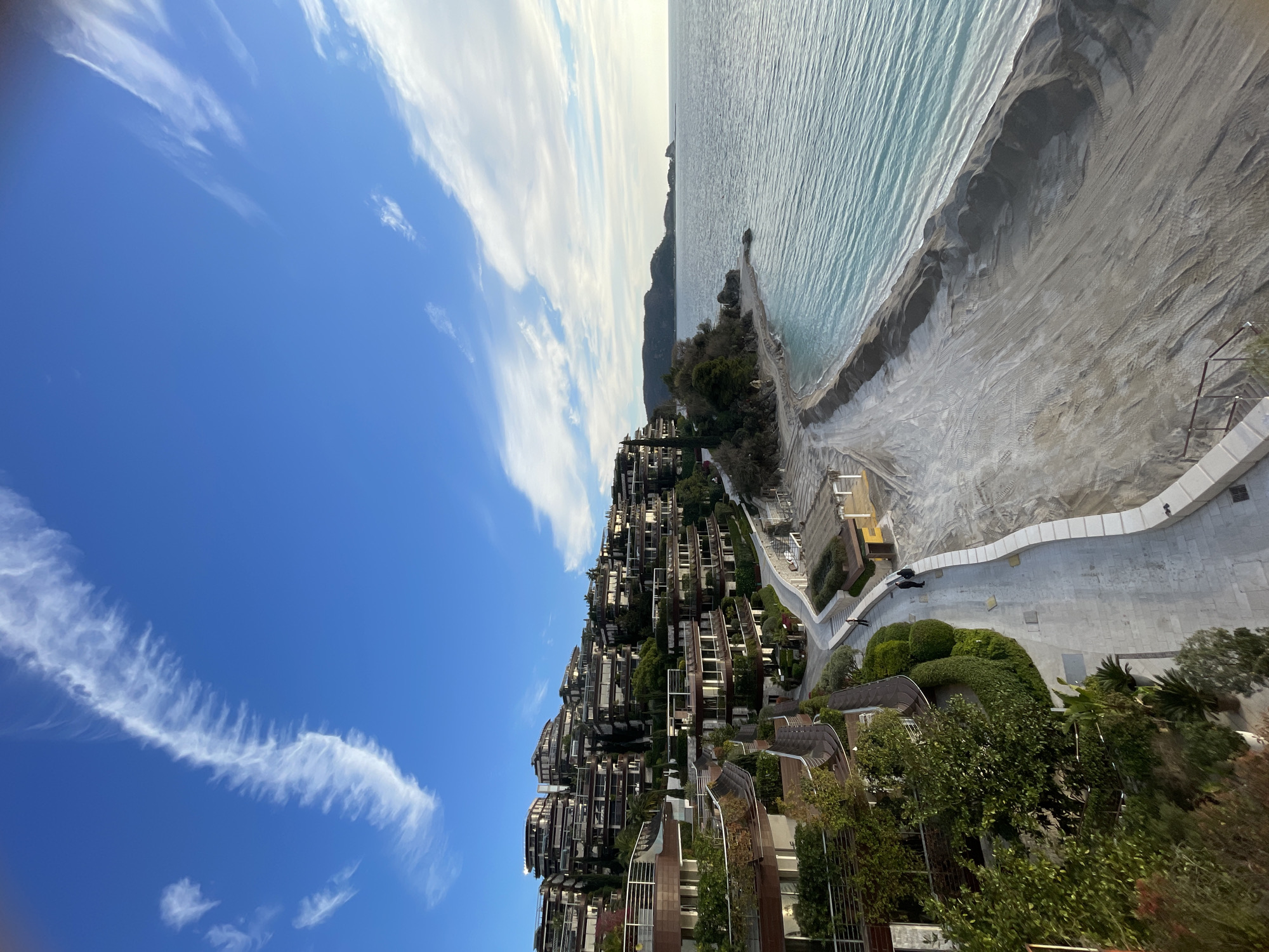 This image depicts a panoramic coastal scene located near the Dukley Gardens complex in Budva, Montenegro.
The upper third of the frame shows a clear blue sky with scattered white cirrus and cirrocumulus cloud formations, including a prominent, elongated, feathery cloud arching from the upper left towards the center.
The right half of the image features a calm body of turquoise-to-deep-blue water, identified as the Adriatic Sea, with subtle surface ripples. A distant, mountainous headland or island is visible on the horizon towards the right.
In the foreground and extending along the coast, a wide, light-colored sandy beach occupies the lower right quadrant. The sand exhibits numerous tire tracks and linear formations, indicative of recent disturbance or renourishment. Several indistinct, light-colored beach structures, possibly collapsed or partially erected shade devices, are present on the sand near the walkway.
The left half of the image is dominated by numerous multi-story, terraced buildings, consistent with the Dukley Gardens complex. These structures feature light-toned facades, dark-framed windows, and extensive balconies with glass or metal railings. Many levels incorporate lush green vegetation as part of integrated terrace gardens. A paved walkway, constructed of light-colored stone or concrete, meanders horizontally across the mid-ground, paralleling the coastline and separating the buildings from the beach. This walkway is bordered on the beach side by a low, curved white retaining wall. Lush green vegetation, including trees and manicured shrubs, is abundant on the hillsides and terraced levels.
Five human figures are present on the paved walkway. Two individuals are walking together towards the right in the middle-ground. One solitary individual is further along the path, also walking away from the viewer. Two additional individuals are standing close to the retaining wall, observing the sea or interacting.
The scene is illuminated by natural light, suggesting either early morning or late afternoon based on the soft quality of light and the presence of elongated shadows.