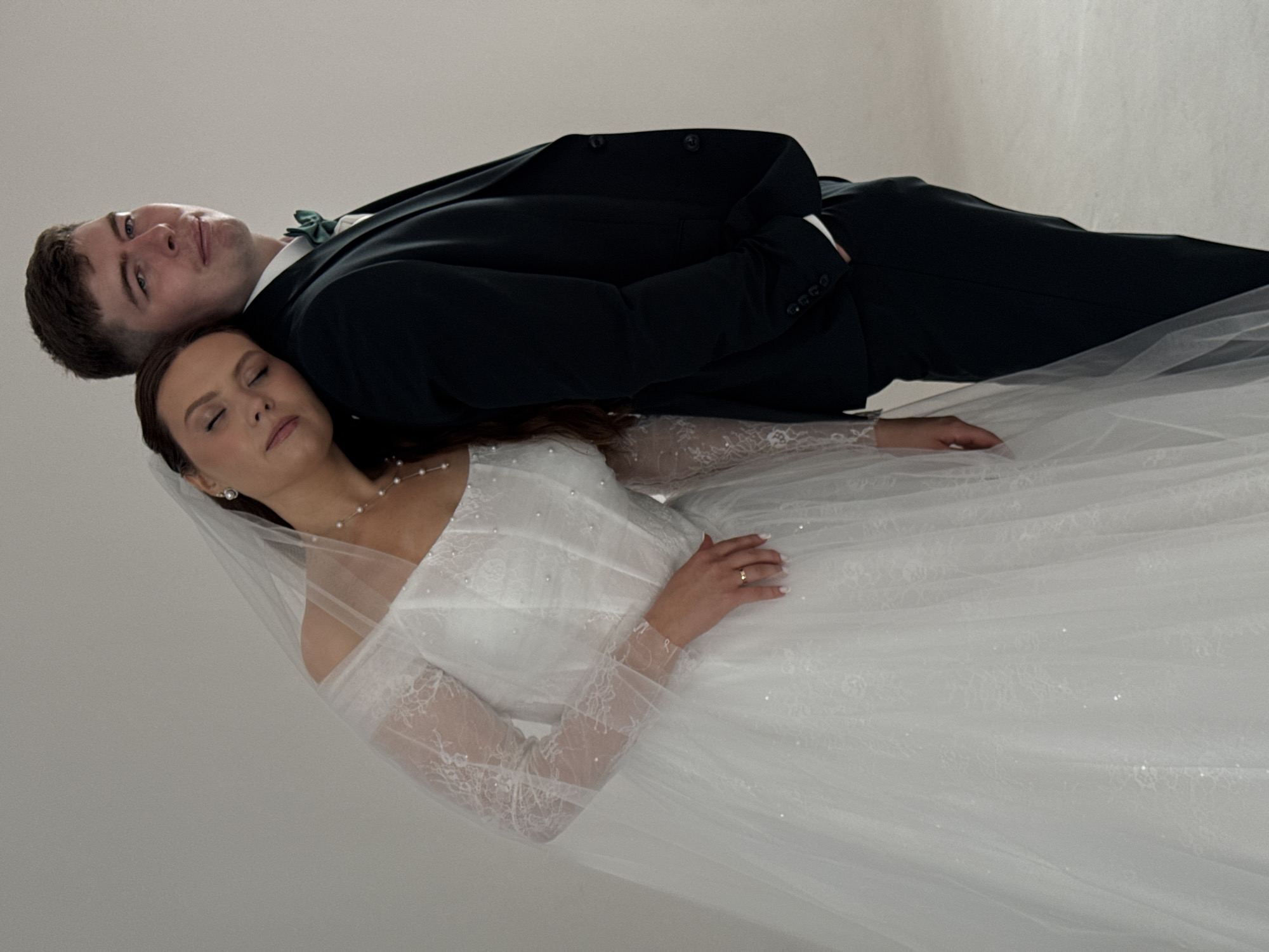 A full-shot image captures a wedding couple positioned horizontally against a plain, light grey background, indicative of an indoor studio setting. The man is on top, wearing a dark suit with a white collared shirt and a green bowtie. He has short, dark hair and is looking directly at the camera with a subtle, content smile. Below him, the woman is resting her head on his shoulder or upper arm. She is adorned in a white wedding gown featuring long, sheer lace sleeves, a pearl necklace, and pearl earrings, with a delicate veil draped behind her head. Her eyes are closed, conveying a serene and peaceful expression. Her right hand, adorned with a wedding ring, rests gently on the fabric of her dress. The couple's bodies extend towards the right of the frame, with the voluminous white skirt of the woman's gown filling the lower right portion of the image. The overall lighting is soft and even. The image depicts an intimate moment between the newlyweds during what appears to be a wedding photoshoot. The location context is Chaikovsky, Russia, though no specific geographical cues are visible in the frame. There is no visible text.