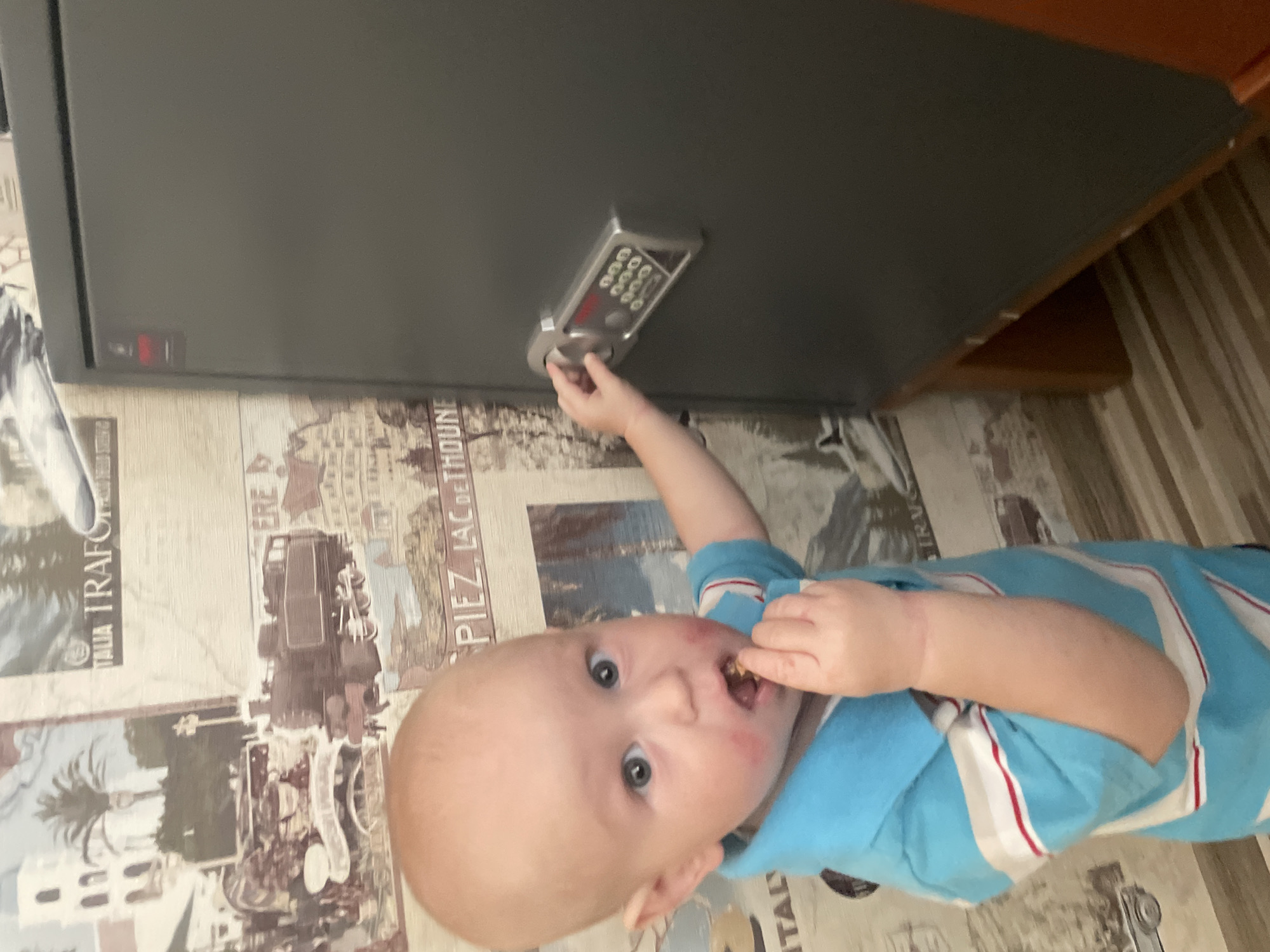 A fair-skinned toddler, with fair hair and blue eyes, is standing in front of a dark gray safe. The toddler is wearing a blue collared shirt with white and red stripes on the sleeves. The toddler is eating something held in their right hand. The toddler's face has red smudges around the mouth. The toddler's left hand is reaching out towards the safe's keypad. The background consists of wallpaper depicting vintage travel posters and trains. The floor is made of wooden planks. The location is in the city of Novyi Nalim, Russia.