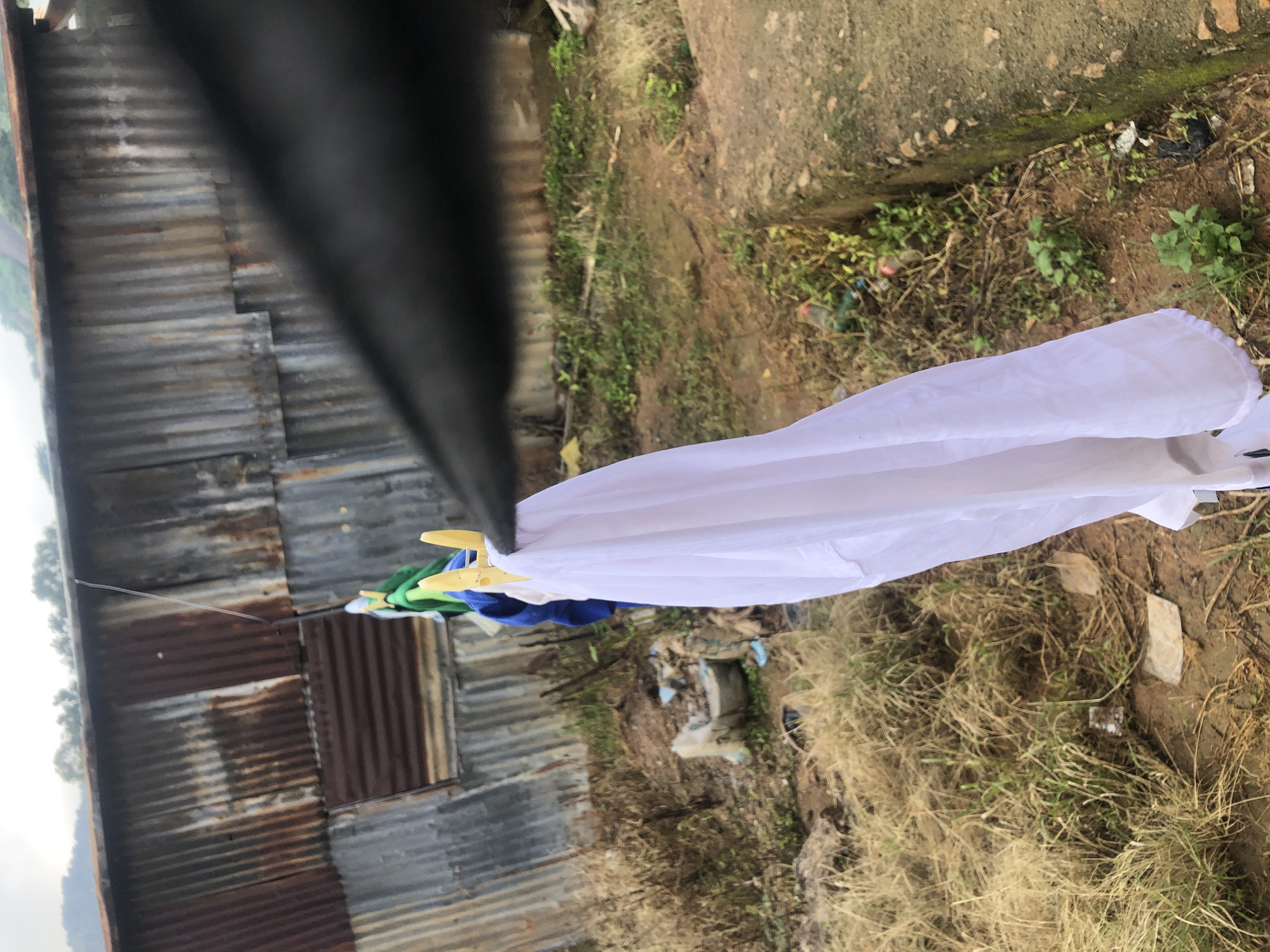 In Jos, Nigeria, a close-up, eye-level shot captures laundry hanging on a clothesline against a backdrop of a weathered, corrugated metal structure. The metal sheeting exhibits significant rust and discoloration, indicating age and exposure to the elements. A portion of a dark fabric, possibly an umbrella or garment, is prominently in the foreground, partially obscuring the view. Several pieces of clothing, including blue and green fabrics, are clipped to the line with yellow clothespins. A large white cloth hangs prominently in the center, its fabric gently draped. The ground in the foreground is covered with dry, yellowish grass and dirt, with some scattered debris and vegetation. The overall impression is one of an outdoor laundry scene in a somewhat unkempt environment, likely a residential area in Jos.