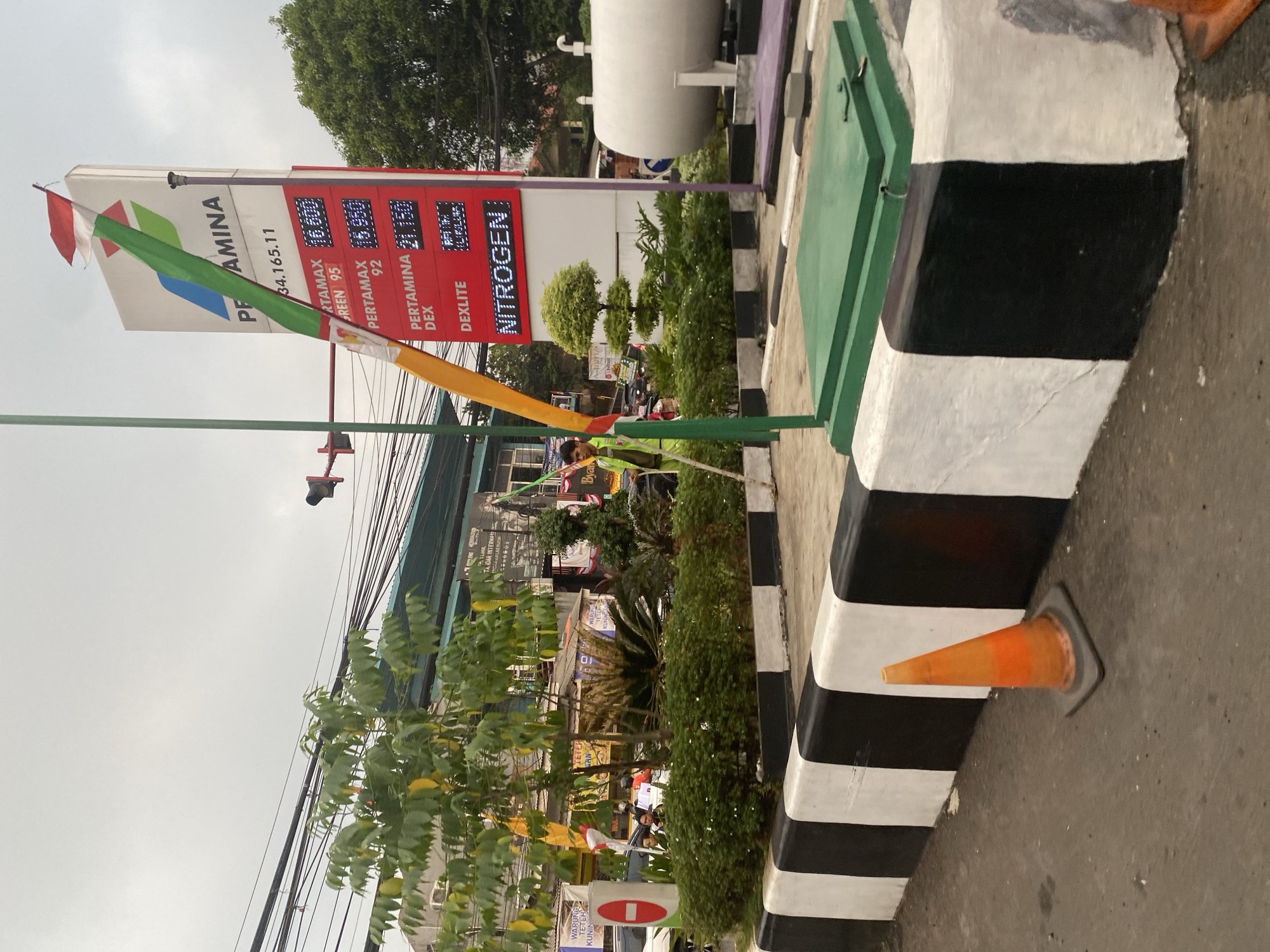 The image depicts a Pertamina gas station in Depok, Indonesia. A prominent Pertamina sign displays fuel prices: Pertamax 95 at 16,900, Pertamax 92 at 16,950, Pertamina Dex at 21,150, and Dexlite. The sign also indicates a phone number, 34.165.11. A young man in a bright green uniform is visible near the sign, and a large white gas tank is to the right. The gas station features a decorative border with black and white stripes along the ground. An orange traffic cone is positioned on the pavement in the foreground. Lush green bushes and trees surround the area, with power lines and buildings in the background. The sky is overcast.