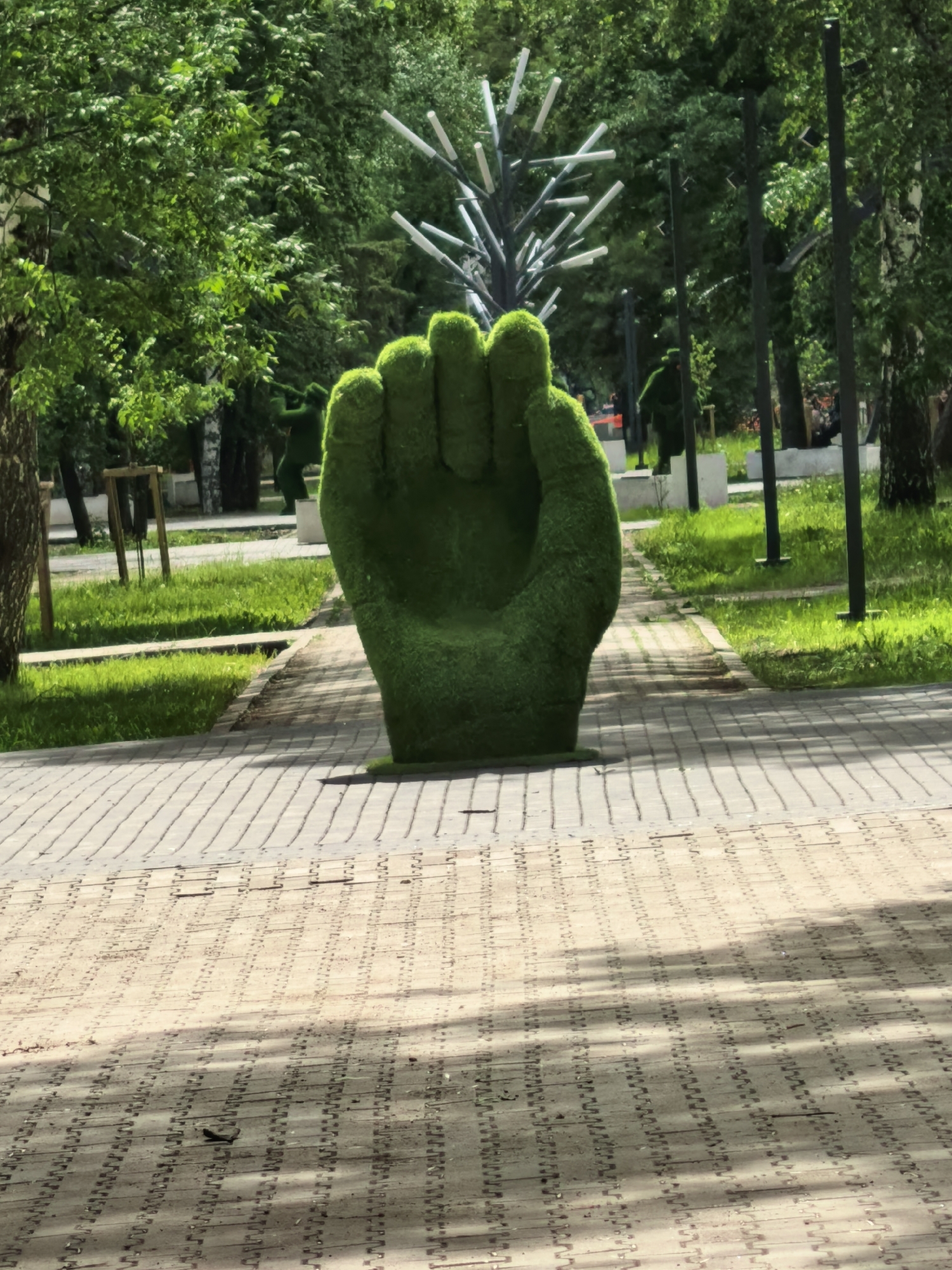 A ground-level full shot depicts a large, sculpted green fist placed on a brick walkway in what appears to be a city park in Sterlitamak, Russia. The fist, resembling a grass topiary, is the focal point. The walkway consists of light and dark gray bricks laid in a uniform pattern. Behind the fist, there is a patch of green grass and other trees. A metal structure with white horizontal elements is seen further back. In the background, there are additional green topiary sculptures. Black lamp posts line the walkway behind the fist. Green trees provide a backdrop.