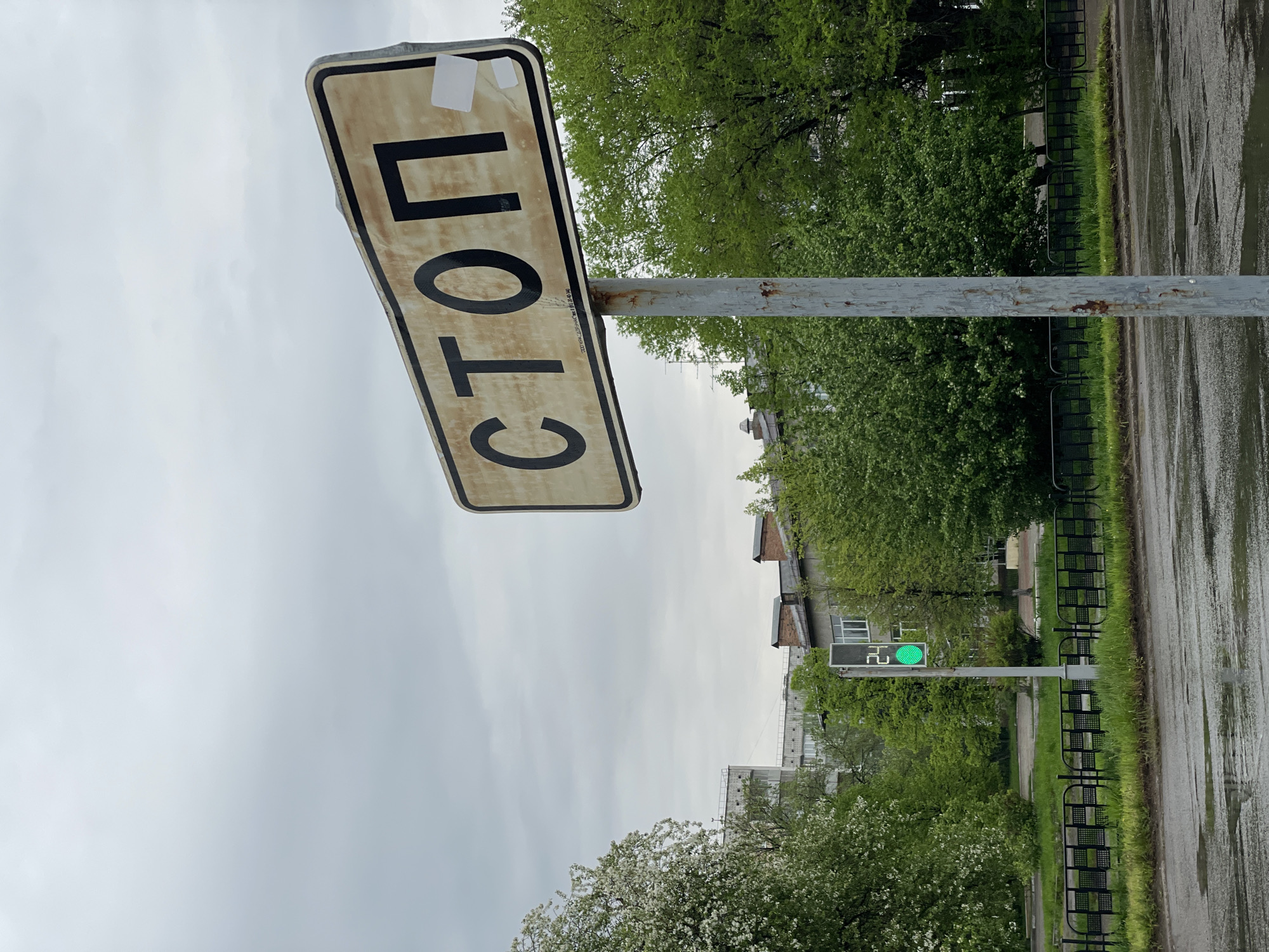 The image shows a street scene in Zheleznogorsk, Russia, under an overcast sky. A weathered stop sign, reading "СТОП" in Cyrillic, is prominently displayed on a corroded metal pole. In the background, a green traffic light is visible. Puddles on the street indicate recent rain. Trees and a fence line the roadway. Several buildings can be seen in the distance.
