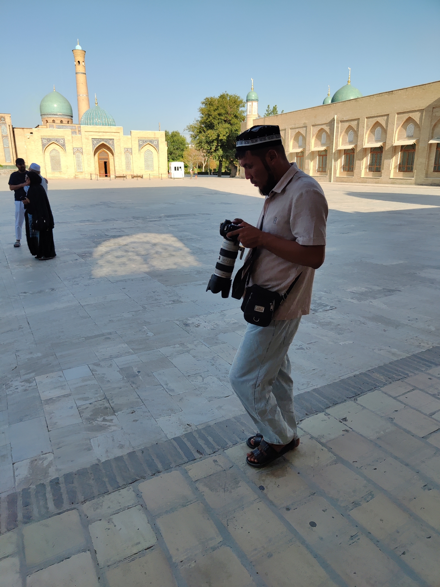 A man wearing a prayer cap and a light-colored shirt is standing in a large, paved courtyard, holding a camera with a telephoto lens. He is looking down at the camera and appears to be adjusting it. He is also wearing light-colored pants and dark sandals, and has a small black bag slung across his body. In the background, there is a large, beige building with multiple green domes and minarets, consistent with Islamic architecture. Two individuals, one in black attire and another in white pants and a dark shirt, are standing to the left of the frame, near the building. The sky is clear and blue, suggesting it is daytime. The paved courtyard is made of large, square stones. The architectural style and the presence of the building suggest this scene is likely located at the Hazrati Imam Complex in Tashkent, Uzbekistan.
