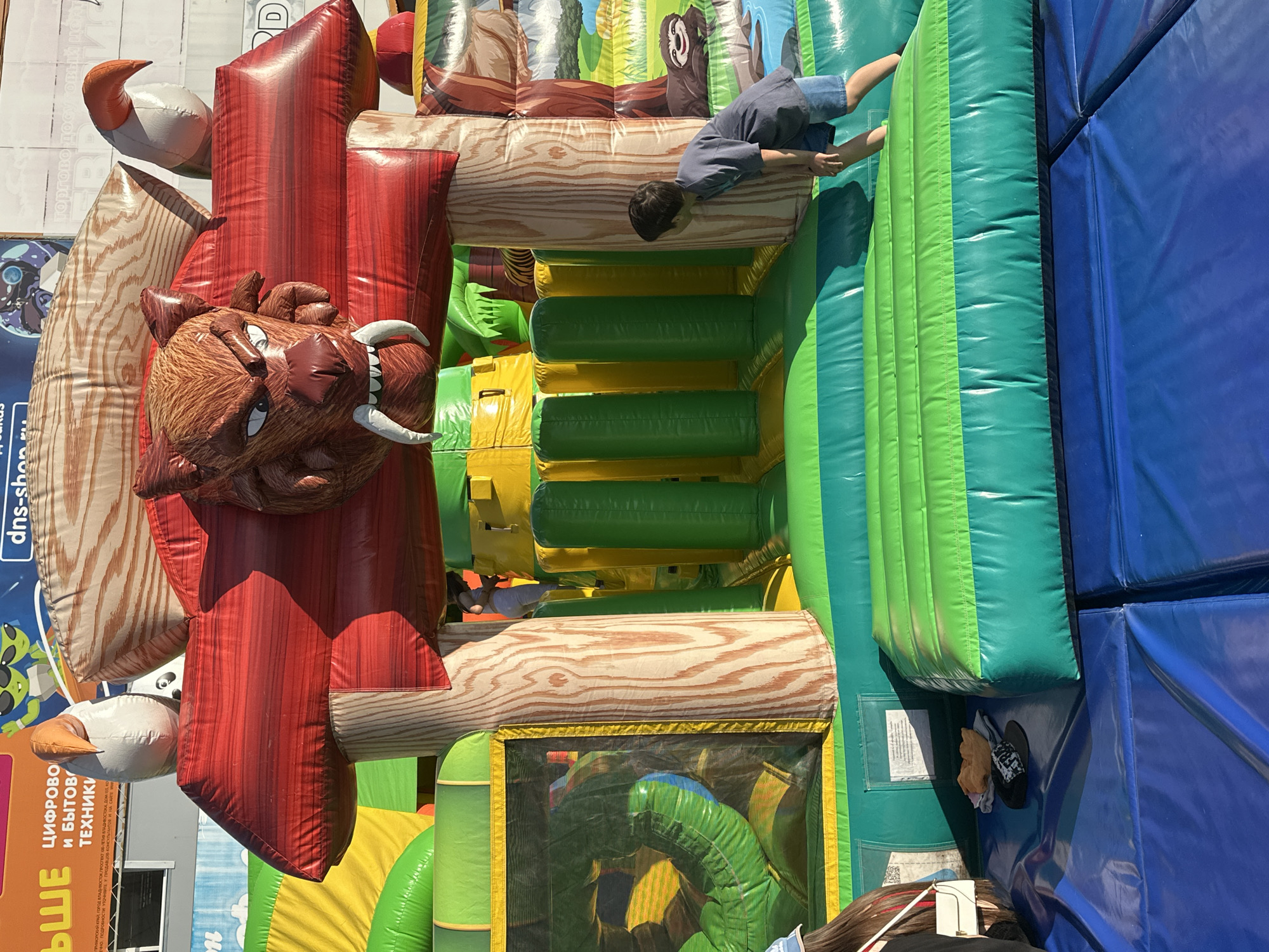 In the city of Uchaly, Russia, a young boy in a blue t-shirt and denim shorts is playing on a large, colorful inflatable obstacle course. The inflatable structure features a prominent, cartoonish tiger head with sharp white fangs at the top, adorned with red and wood-grain patterned panels. The course itself is a mix of green, yellow, and red inflatable elements, with vertical green and yellow columns. A dark mesh window on the left reveals more of the interior structure, a bright green, curved inflatable slide. The boy is positioned on a green, tiered inflatable platform, leaning forward as if about to jump or crawl further into the structure. In the foreground, the blue safety mats of the play area are visible, with a black cap and a folded piece of cloth resting on them. The scene appears to be outdoors, with daylight illuminating the inflatable.
