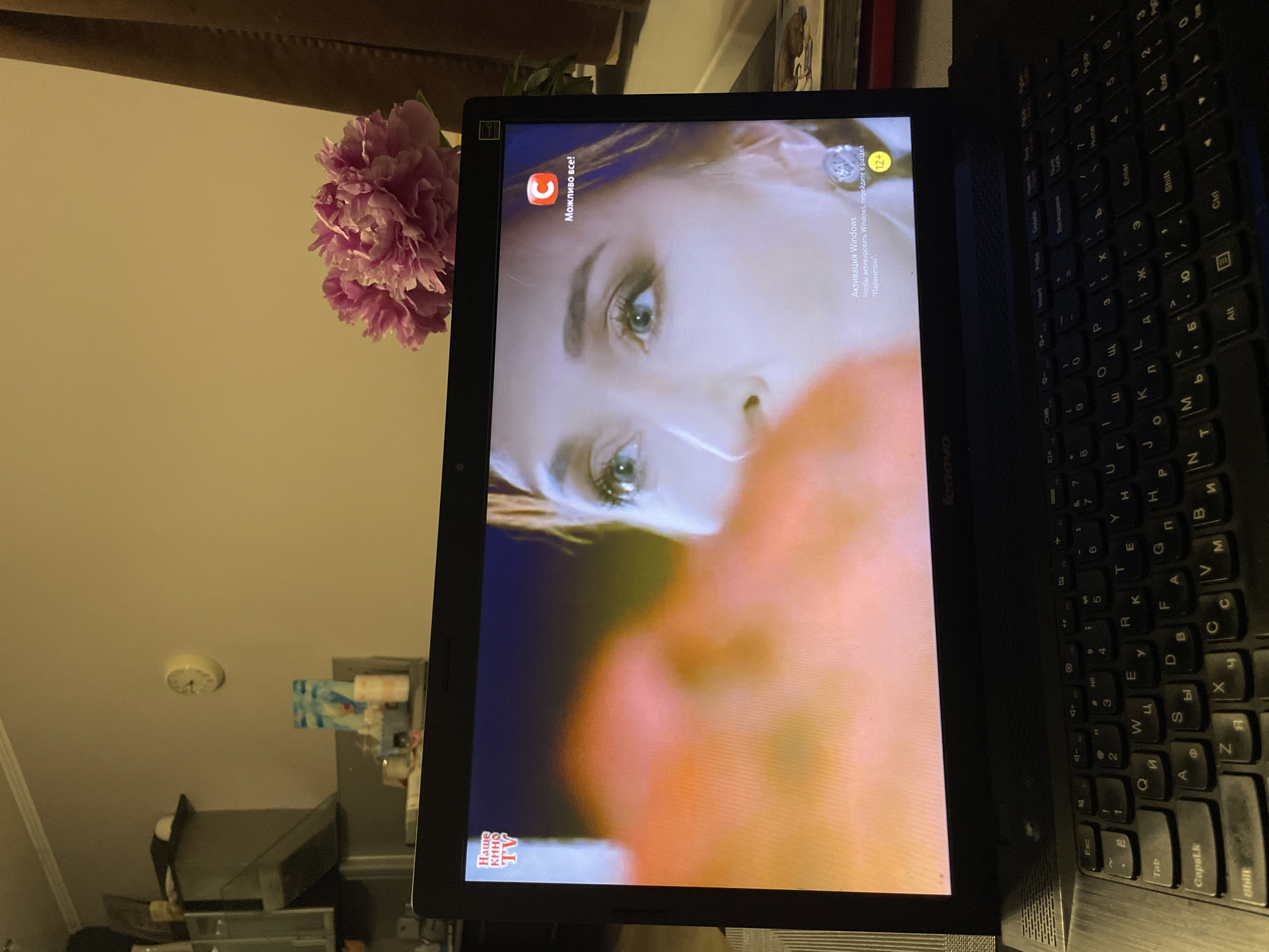 The image displays an indoor domestic scene in Sochi, Russia. A black Lenovo laptop with a Cyrillic keyboard is open in the foreground, positioned on a textured surface. The laptop screen displays a close-up shot of a woman with light-colored eyes (appears green or blue) and light brown/reddish hair, looking upwards or to the right. In the top left corner of the screen, the logo "НАШЕ КИНО TV" (Our Cinema TV) is visible. The top right corner features a red logo with a white "C" and the text "Можливо все!" (Everything is possible!). A "12+" age rating icon is present in the bottom right corner of the screen, above a Windows activation watermark that reads "Активация Windows / Чтобы активировать Windows, перейдите в раздел "Параметры"" (Activate Windows / To activate Windows, go to "Settings"). To the right of the laptop, a bouquet of pink peonies is visible. The background consists of a light-colored wall with a small, round analog wall clock mounted centrally, indicating a time between 9:50 and 10:20. To the left, elements of a kitchen or utility area are visible, including a range hood, glass-fronted cabinets, and various objects on a counter. A dark brown curtain or drape is partially visible on the far right. The overall lighting is dim. No other individuals are present in the scene.