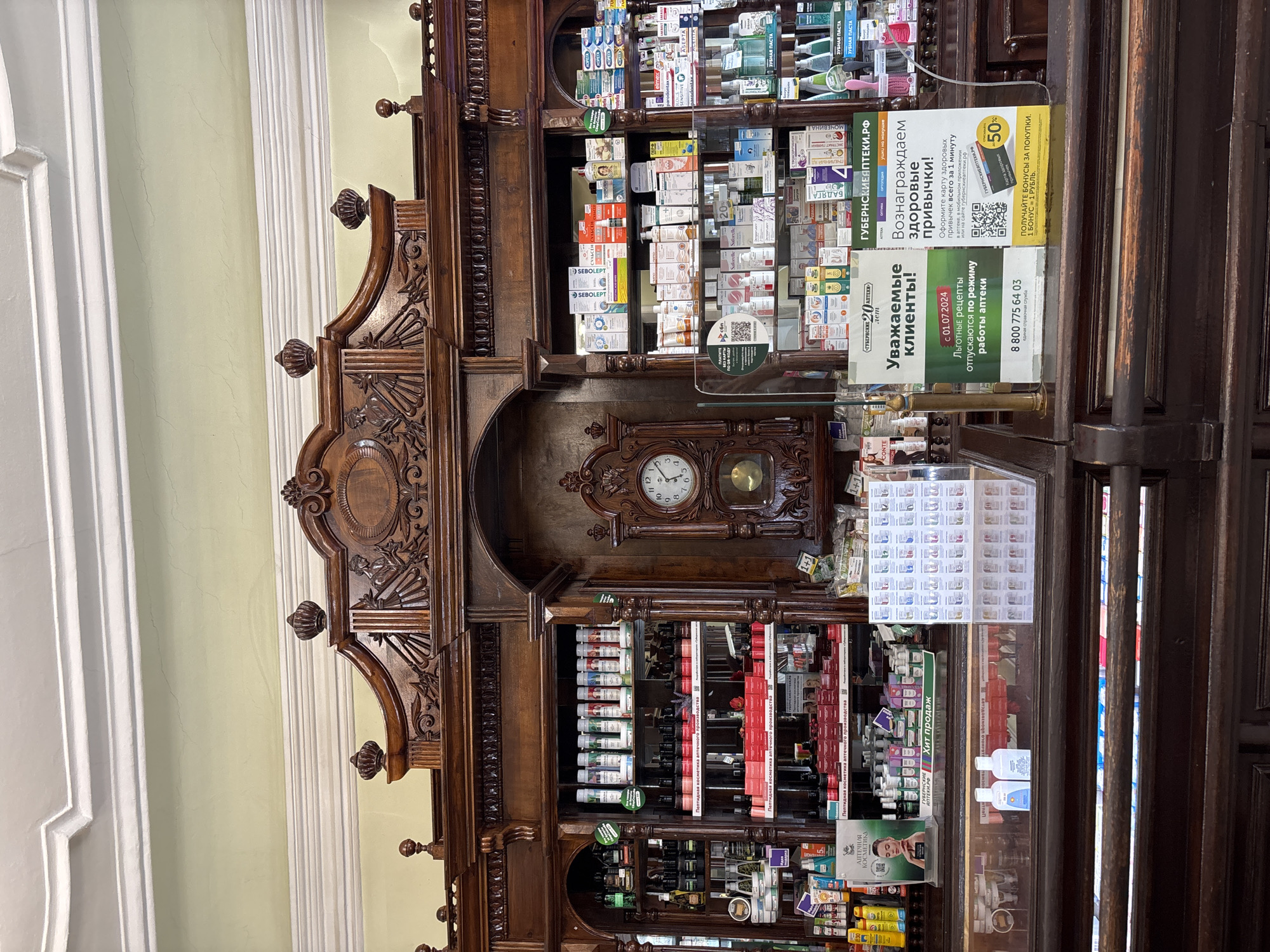 The image depicts the interior of a pharmacy, likely in Krasnoyarsk, Russia. The scene features an antique wooden cabinet filled with various pharmaceuticals. Above the cabinet is an ornate clock. Signage in Russian promotes health rewards, announces the acceptance of preferential prescriptions from July 1, 2024, and displays a customer service phone number. The shelves are stocked with items such as toothpaste, medical cosmetics, and other healthcare products.