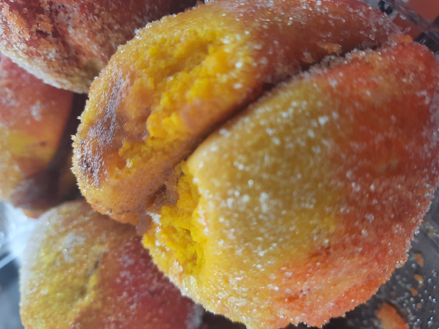 The image presents a close-up, high-angle view of multiple dessert items, likely sweet pastries or confections. The primary subjects are several roundish, peach-colored treats, generously coated in sparkling white granulated sugar.
One prominent item in the foreground and center is split open, revealing a vibrant, soft, and somewhat crumbly yellow interior, suggesting a cake-like or sponge texture. The exterior of these items displays a gradient of colors from warm yellow to orange, with some areas having a slightly darker, reddish-brown hue, possibly from baking or a flavored coating. The sugar coating is dense and uniformly applied, creating a rough, crystalline texture that reflects light.
Several other similar items are visible, clustered together in the background and foreground, some partially obscured and out of focus. They maintain the same characteristic sugar coating and varied coloration. The lighting emphasizes the individual sugar crystals, making them stand out.
No human activity is depicted. The scene focuses entirely on the prepared food items, which resemble "pesche dolci" (sweet peaches), an Italian dessert often made from two halves of cake or cookie dough filled with cream and soaked in liqueur, then coated in sugar. There are no discernible location cues, time of day indicators, or visible text. The overall impression is one of appealing, freshly made sweets.