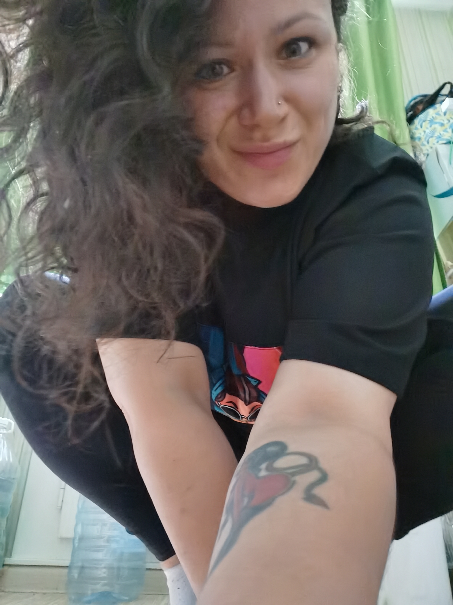 A woman with dark, curly hair is captured in a selfie. She is wearing a black t-shirt and black leggings. A tattoo is visible on her left forearm. She is indoors, and a green curtain is visible in the background. Two large plastic water bottles are on the floor to her left. The environment suggests a residential setting, possibly an apartment in the city of Chaikovsky, Russia.