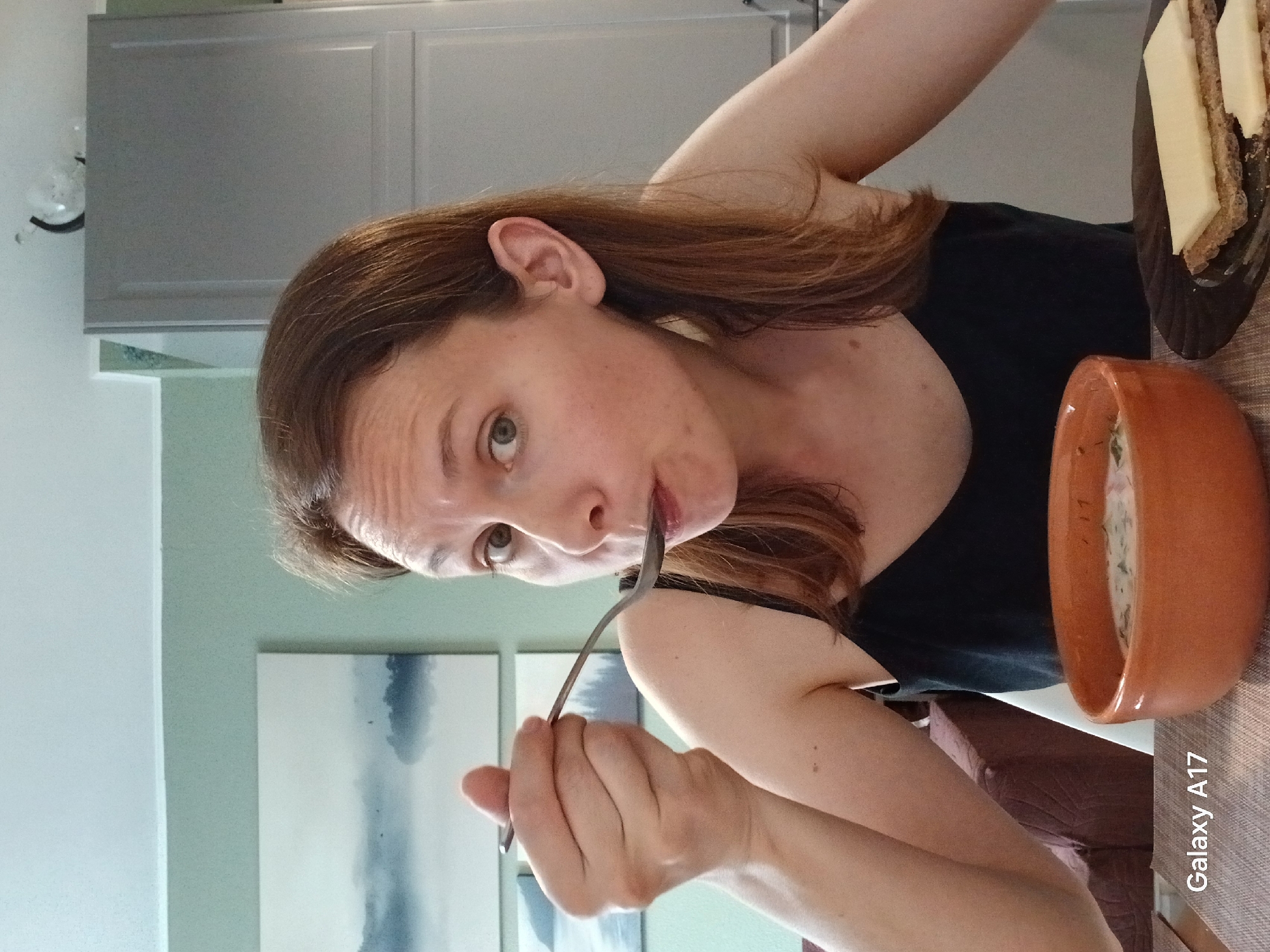 A woman with brown hair is eating something with a fork. There is a brown bowl next to her, containing what looks like soup. She is wearing a black top. Next to the bowl is a plate with some bread topped with a white substance. The background includes a white wall with an abstract painting and kitchen cabinets. There is a "Galaxy A17" watermark in the lower right corner. The location is not discernible from the image.