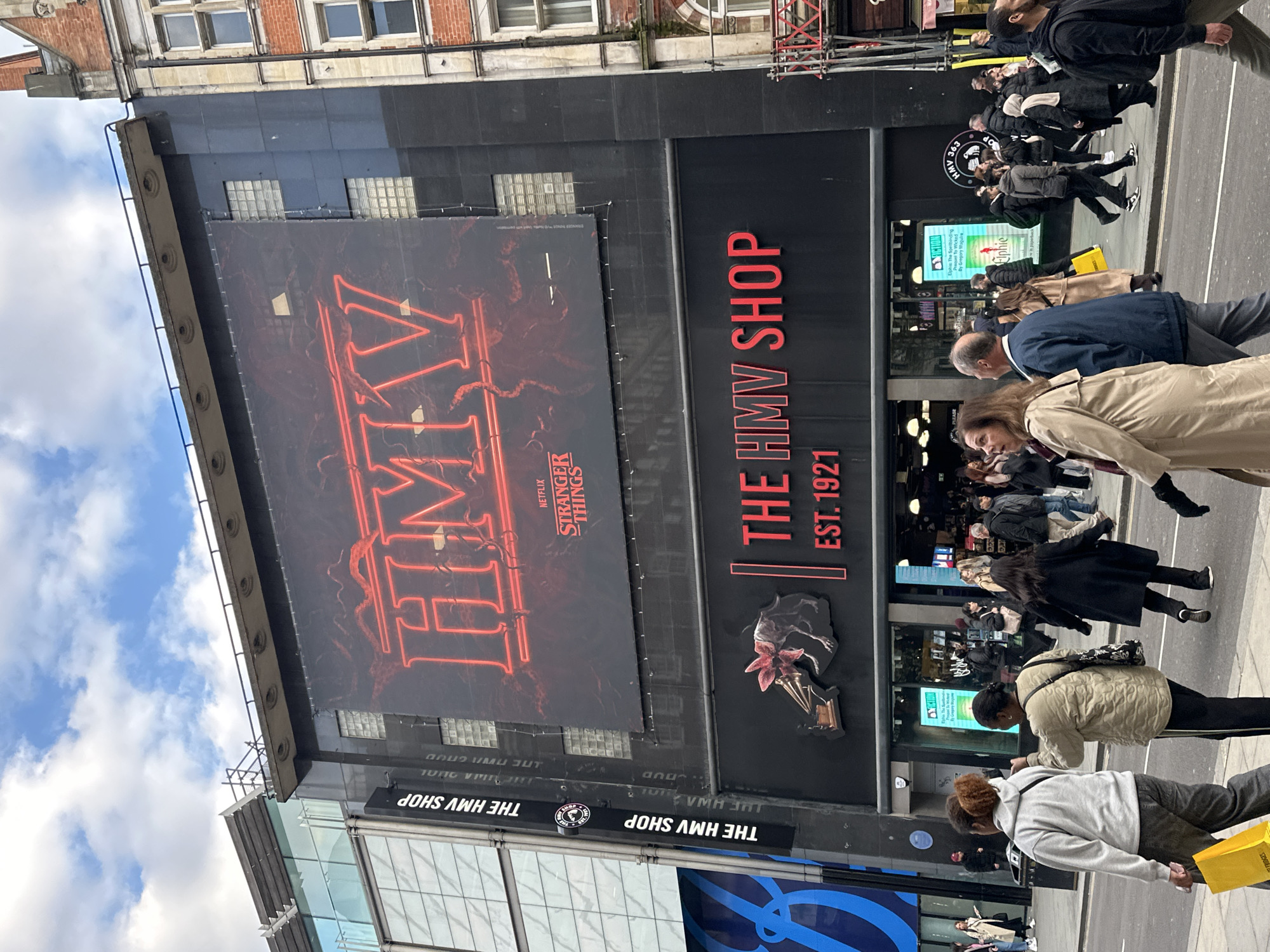 The image shows the exterior of an HMV shop in London, United Kingdom. A large advertisement for the Netflix series "Stranger Things" is displayed prominently on the building. The sign reads "HMV" vertically, with "Netflix Stranger Things" beneath. Beside the advertisement, a sign reads "THE HMV SHOP EST. 1921." People are walking on the sidewalk in front of the shop. Some are looking at the shop while others are walking past. The sky is partially cloudy. The building is mixed between glass and brick.
