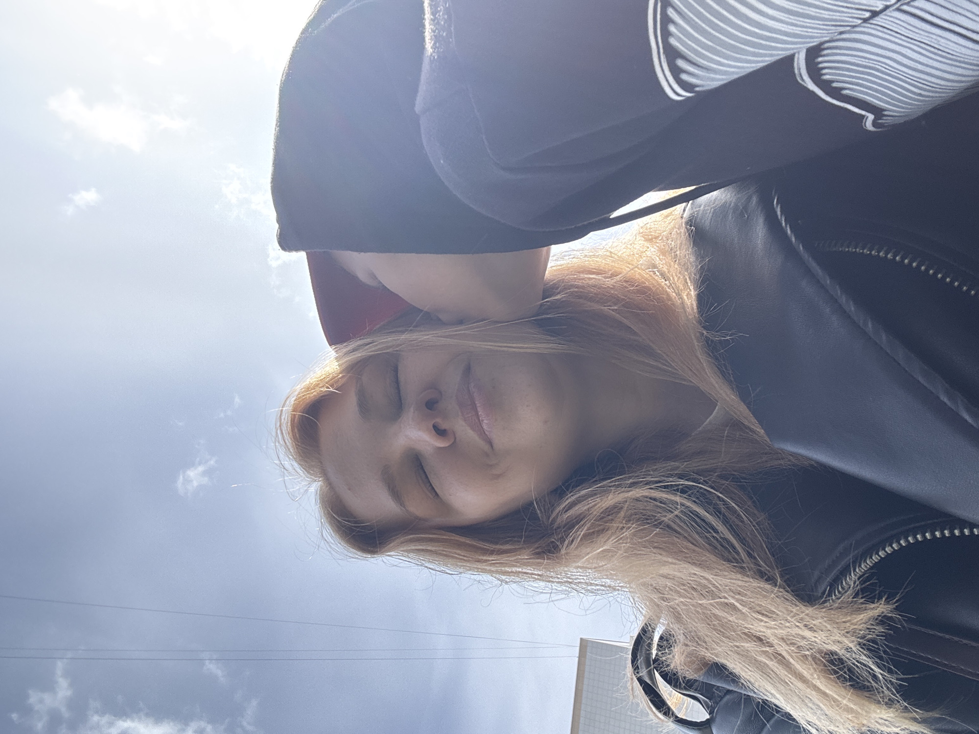 A selfie is taken from a low angle, with the subject's face tilted upwards. The background shows a cloudy sky and the top of a building, possibly indicating an outdoor urban setting. The subject has light-colored hair and closed eyes. A second person is partially visible in the upper portion of the frame, wearing a black hoodie and a red baseball cap. The subject also has on a black jacket with visible zippers. The location is Rusanovo, Russia.