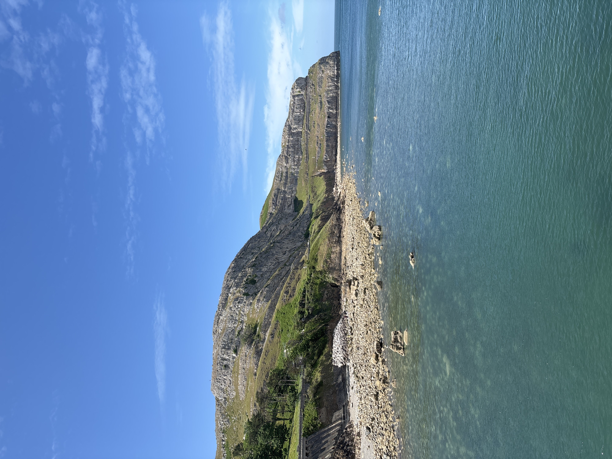 The image captures a scenic view of the Great Orme, a headland on the north coast of Wales, near the town of Llandudno in the United Kingdom. A tall, limestone cliff dominates the center of the image, its face marked by horizontal striations and a patchwork of green vegetation clinging to its slopes. At the base of the cliff, a narrow stretch of rocky coastline transitions into the sea. The water is a pale turquoise color, clear enough to reveal the submerged rocks beneath its surface. The sky is a clear blue with wispy white clouds scattered across it. The image is taken from a low angle, emphasizing the height of the cliff and the expanse of the sea.