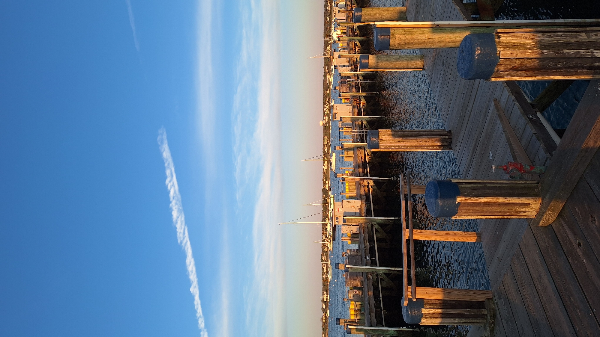 This image presents a serene view of a wooden dock or pier in Nantucket, United States of America, bathed in the warm, golden light of either sunrise or sunset. The scene is dominated by the long stretch of the pier, which extends from the foreground towards the distant horizon, lined with numerous wooden pilings. Many of these pilings are topped with distinctive blue caps or fenders, and the low sun casts long, soft shadows across the weathered wooden planks of the dock.
Along the pier, various boats are moored, predominantly showing the masts of sailboats and the profiles of several motorboats. The calm water of the harbor gently reflects the sky and the dock structures, with subtle ripples visible on its surface. In a closer section of the dock, a red fire hydrant or valve can be seen mounted on a wooden post.
The background features a low, distant shoreline populated with an array of houses and buildings, indicating a coastal settlement. Above the horizon, the sky transitions from a rich, clear blue overhead to a softer, lighter shade near the vanishing point of the horizon. Several wispy clouds are scattered across the sky, along with a prominent, linear contrail stretching diagonally from the upper left. The overall impression is one of peaceful tranquility at a picturesque harbor. No discernible text is visible in the image.