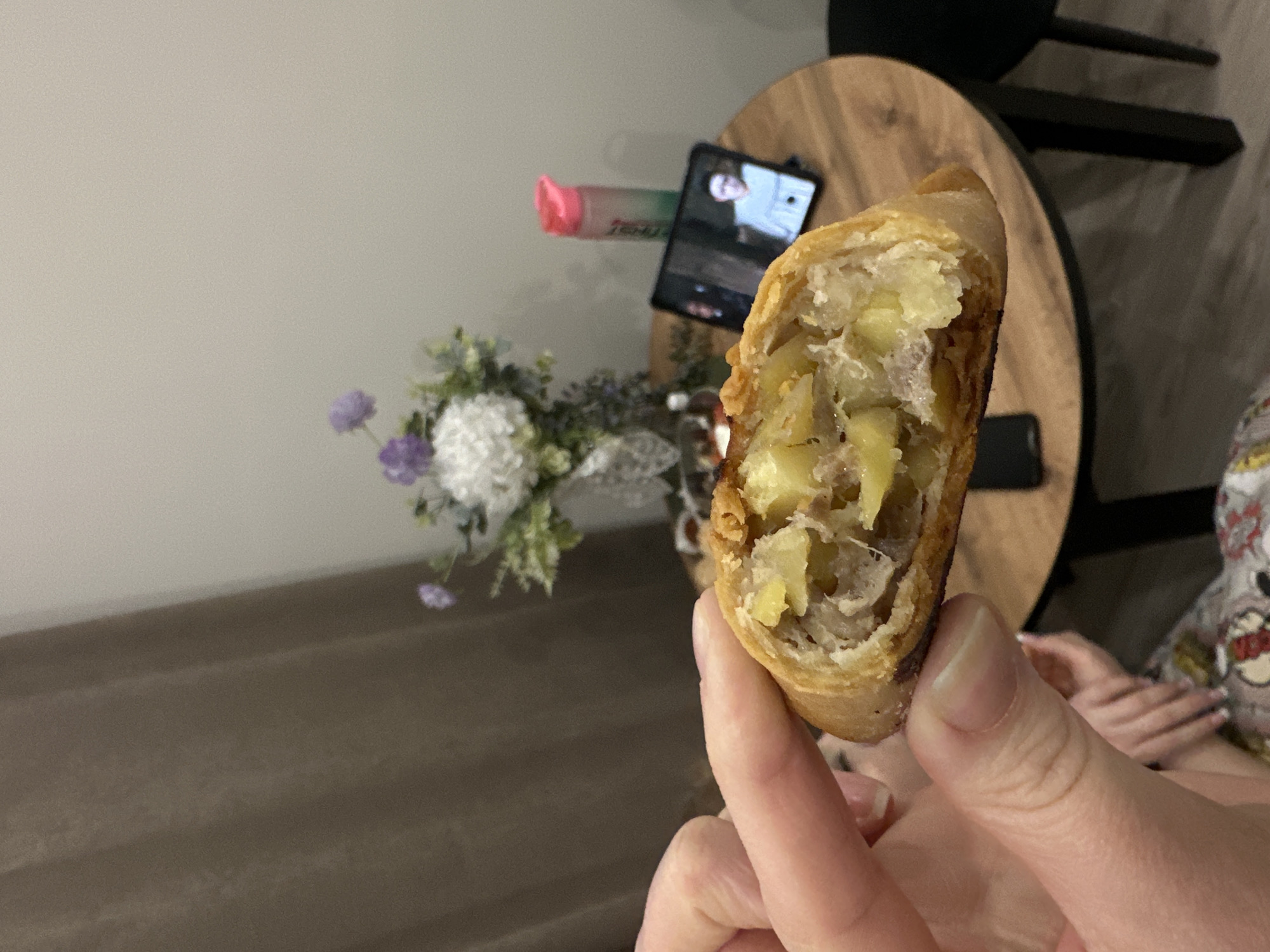 A person holds a cut spring roll, revealing a filling of potatoes and meat. In the background, a round wooden table displays a phone propped up by a pink and green marker. A small bouquet of white and purple flowers sits beside the phone. The location appears to be indoors, likely a residence. The scene does not definitively pinpoint the location to Kazan, Russia.