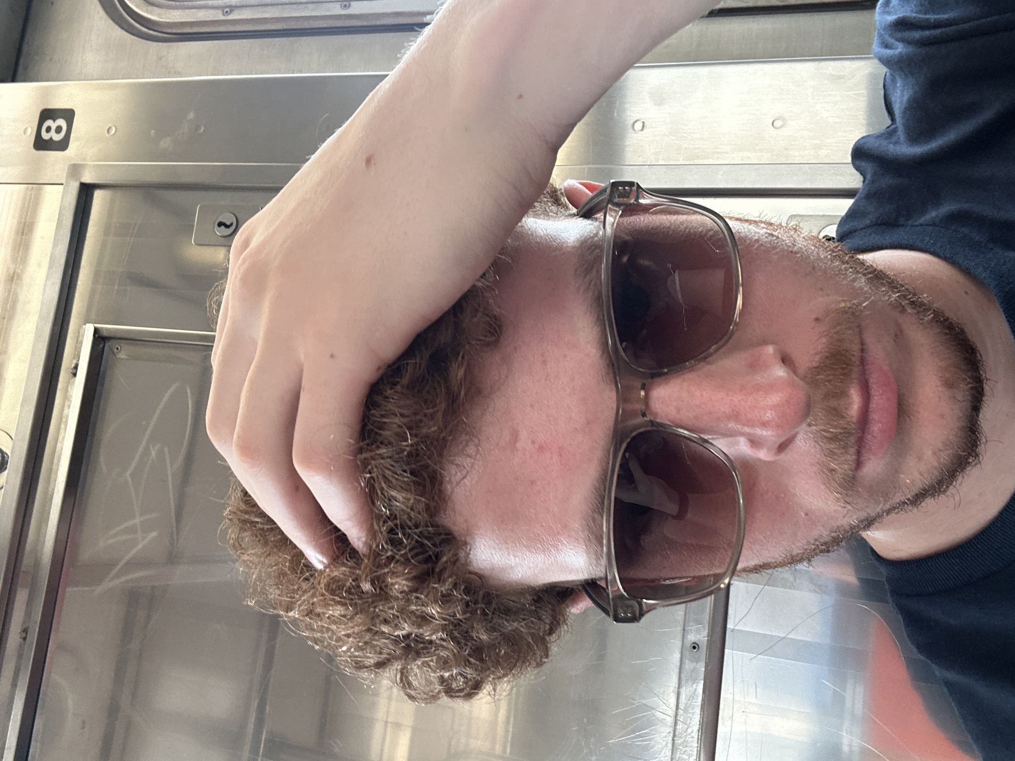 A man with reddish curly hair and a beard is wearing sunglasses. He is inside what appears to be a train car with metal walls. He has one hand on his head. The number "8" is visible on the wall of the train car. The man is likely in New York City, United States of America, as this is a common mode of transportation in the city.