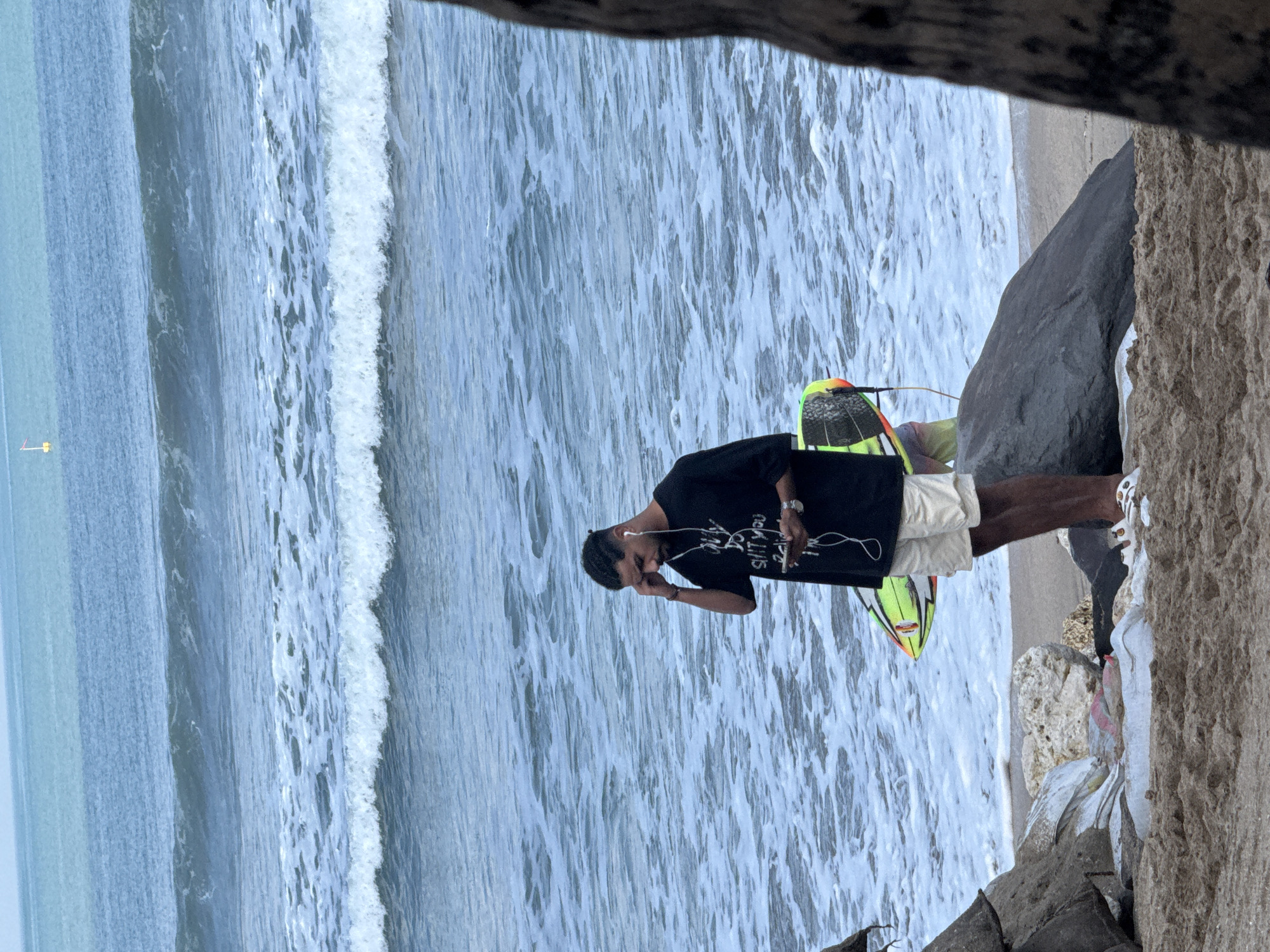 A man stands on the sandy shore of Kuta Beach, Indonesia, near the ocean waves. He is wearing a black t-shirt with text, beige shorts, and white sandals. He holds a smartphone in his right hand and has earbuds in his ears, appearing to be listening to audio. A surfboard with green, yellow, and red accents is propped behind him. The ocean has moderate waves with white foam. In the foreground, large rocks and some debris are scattered on the sand. The horizon shows a light blue sky and a distant yellow marker.