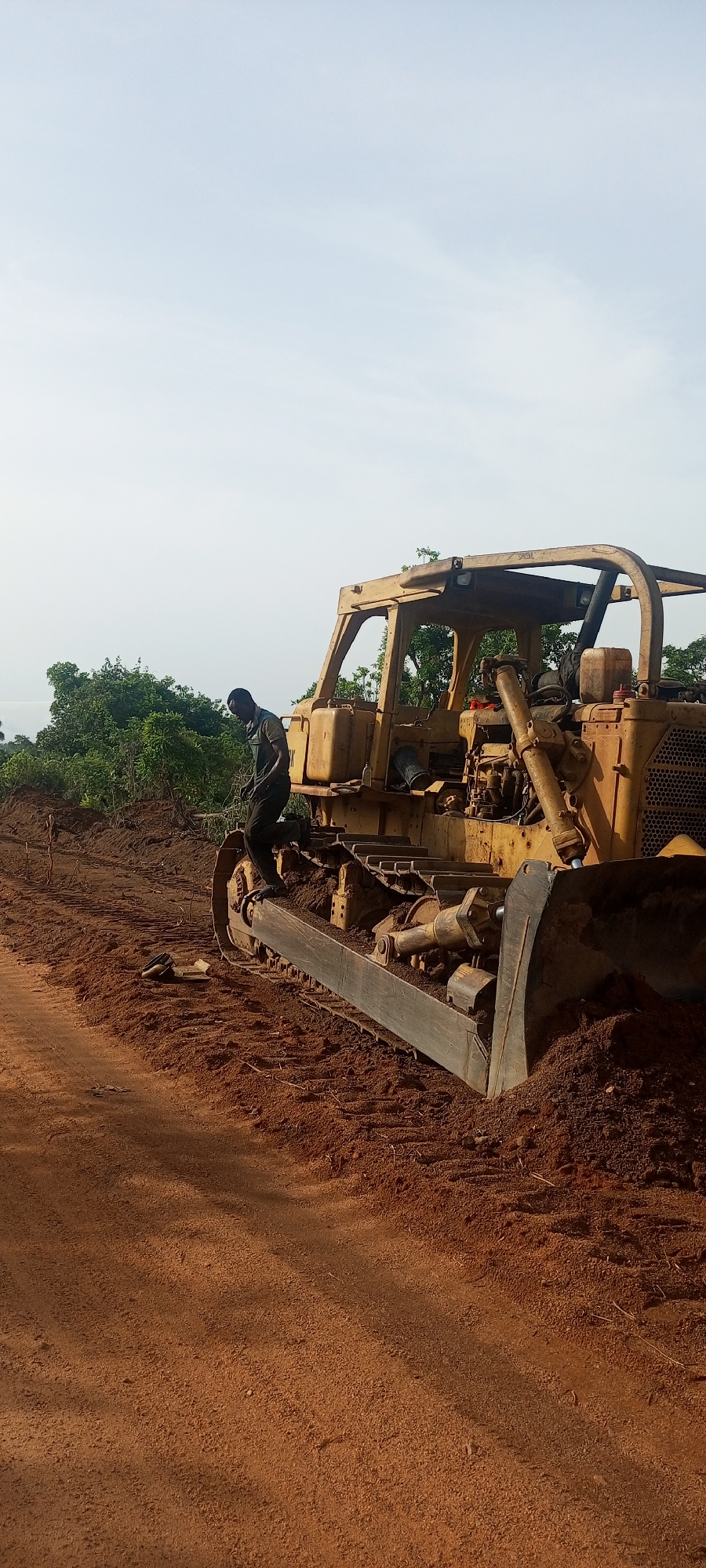 This image captures a scene of land development or construction in Otunade, Nigeria, featuring a large bulldozer and a worker under a clear sky.
The central subject is a robust, yellow crawler bulldozer, appearing well-used with visible wear and tear. It is positioned on a reddish-brown earth surface, characteristic of lateritic soil often found in tropical regions. The bulldozer's large blade is at the front, partially pushed into a mound of earth, suggesting it has either just completed digging or is about to commence operations. Its heavy-duty tracks are also covered in the same reddish-brown soil.
A person, dressed in dark work attire, is standing on the left-side track assembly of the bulldozer. They appear to be focused on the machinery, possibly engaged in an inspection, maintenance, or preparing for operation. Their posture indicates active engagement with the equipment.
The setting is an outdoor, possibly rural or semi-rural landscape. Behind the bulldozer, a dense line of vibrant green trees and bushes stretches across the mid-ground, providing a natural backdrop to the industrious scene. The sky above is a pale, clear blue with very few clouds, indicating bright daylight conditions, likely morning or late afternoon, given the soft illumination without harsh shadows.
The foreground is dominated by the reddish-brown earth, which forms a path or cleared area extending towards the left of the frame. The overall impression is one of ongoing work in a natural environment, utilizing heavy machinery for earthmoving or land preparation. No discernible text is clearly visible on the equipment or in the environment.