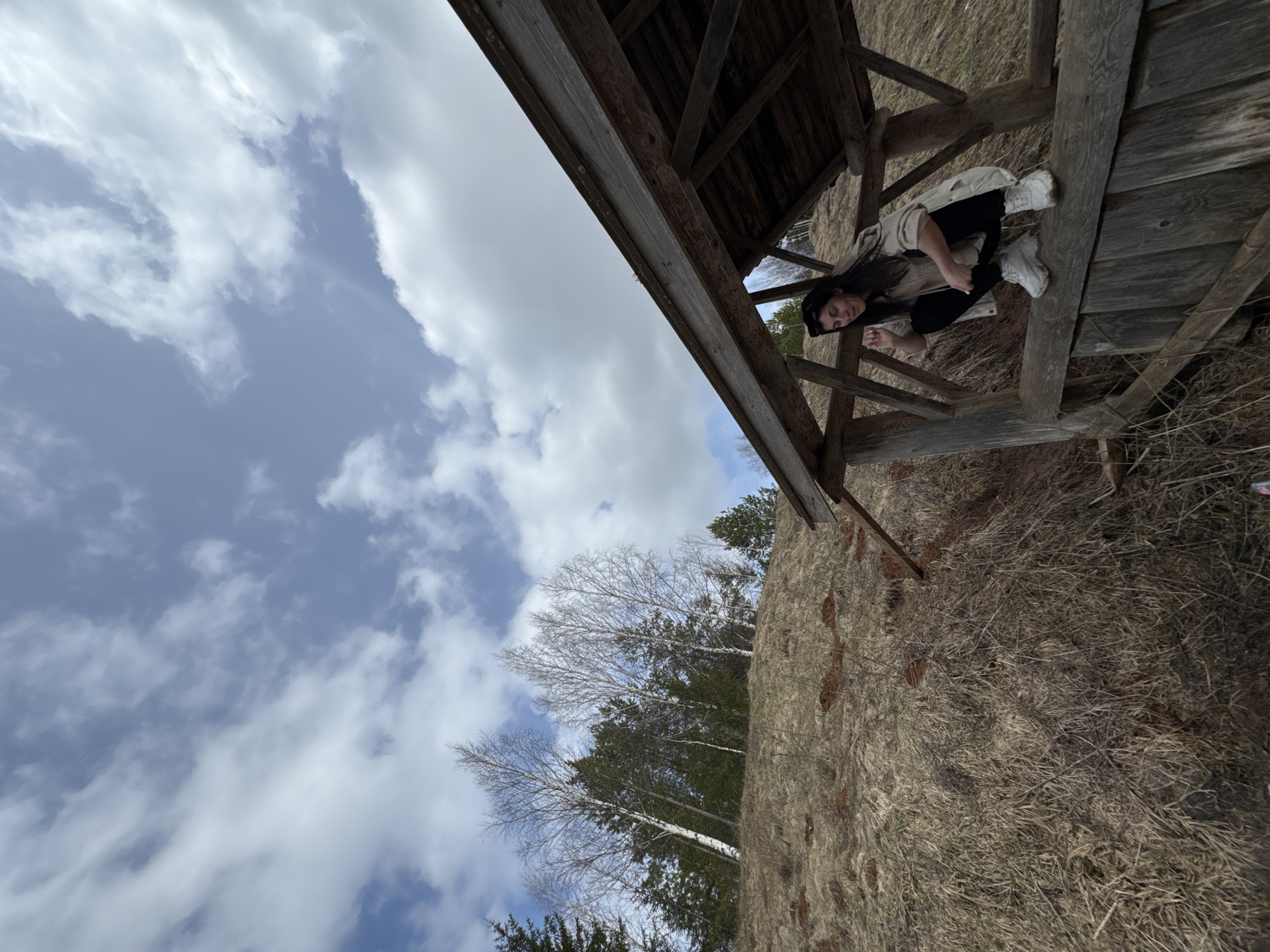 The image depicts a woman sitting on a wooden structure in Вассята, Russia. She is wearing black pants, a beige top, a light-colored jacket, and white sneakers. The wooden structure appears to be a small shelter or platform. The background includes a landscape with dry grass and trees, and a sky with clouds. The image is rotated, with the sky appearing to be on the left side.