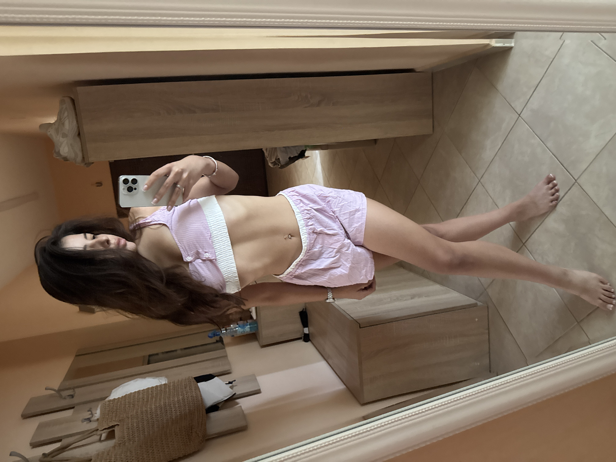A young woman, with long, dark hair, is taking a mirror selfie in what appears to be a bedroom or dressing area. She is wearing a pink and white striped two-piece loungewear set, consisting of a cropped top and shorts. The set has white trim. She has a belly button piercing. In the background, a wardrobe with a light wood finish is visible, with a pair of sneakers on top. There is also a dresser with a dark wood finish. A straw bag hangs on a hook on the wall. The floor is tiled. The lighting suggests daytime. The image is taken indoors and the environment appears to be domestic. There are no discernible elements that place the scene specifically in the city of Plovdiv, Bulgaria.