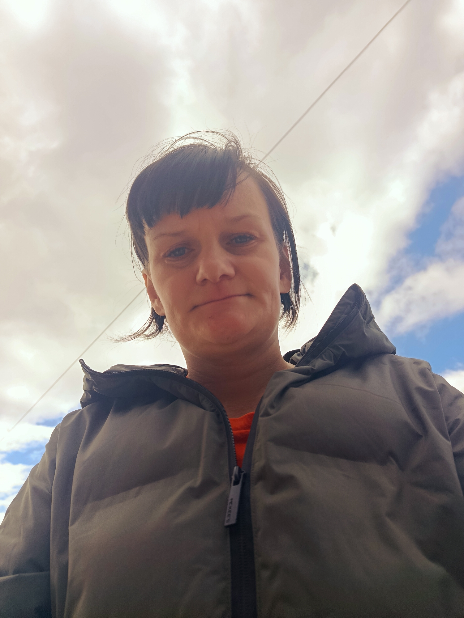 A low-angle selfie of a woman with short, dark hair and bangs. She is wearing a gray puffer jacket over an orange shirt. The jacket is zipped up. The sky is visible in the background, with a mix of clouds and blue sky. A wire or power line is visible in the upper part of the image. The photograph was taken in Фоки, Russia.