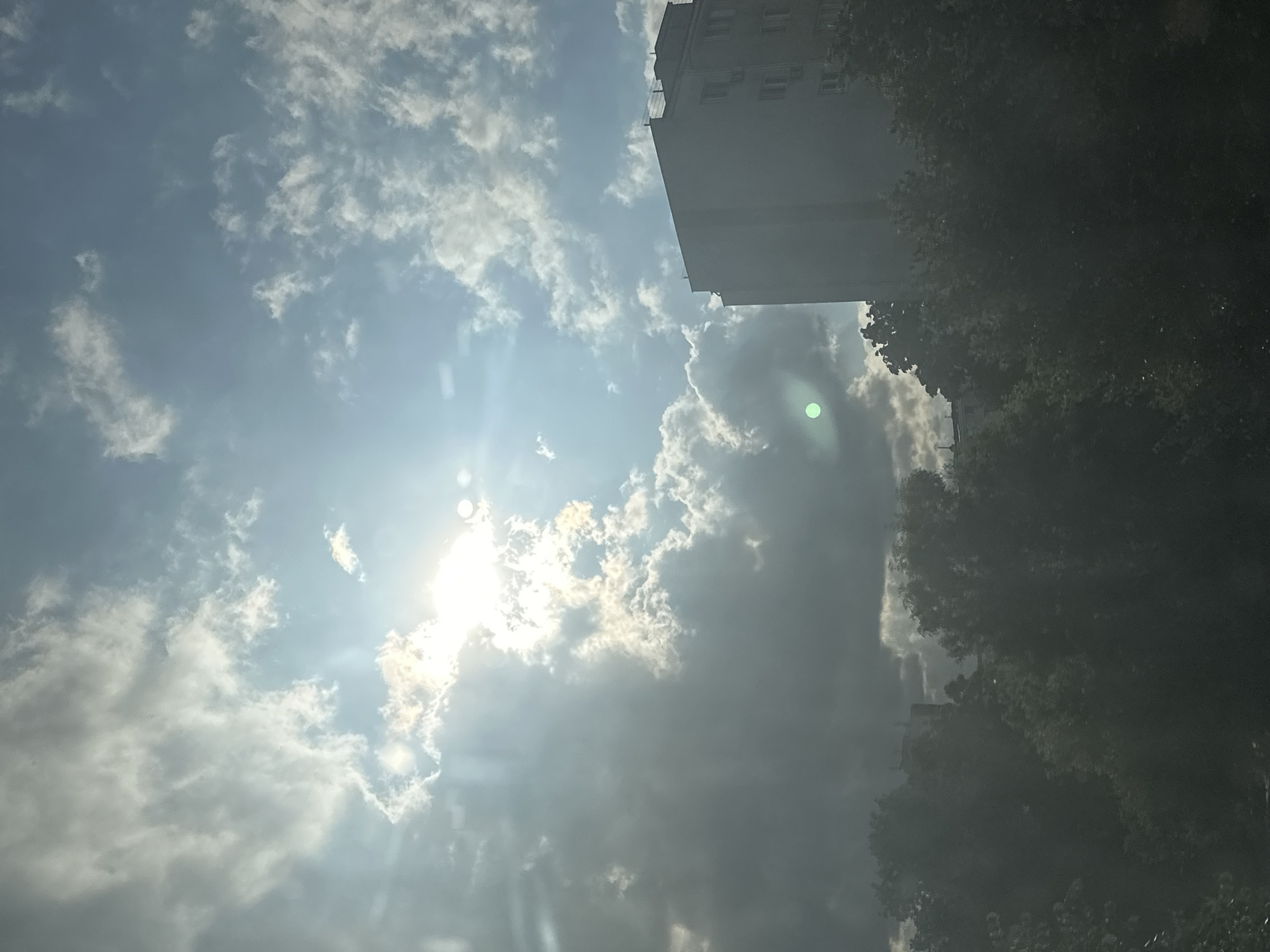 The image captures a cloudy sky with the sun peeking through, creating lens flare effects. A tall, light-colored building stands on the right side of the frame, partially obscured by trees in the foreground. The trees are dark and dense, covering the lower portion of the image and extending to the left. The sky is a mix of bright blue and heavy, gray clouds. A bright green circular anomaly is visible in the sky, to the left of the building. The overall scene suggests an outdoor urban environment.