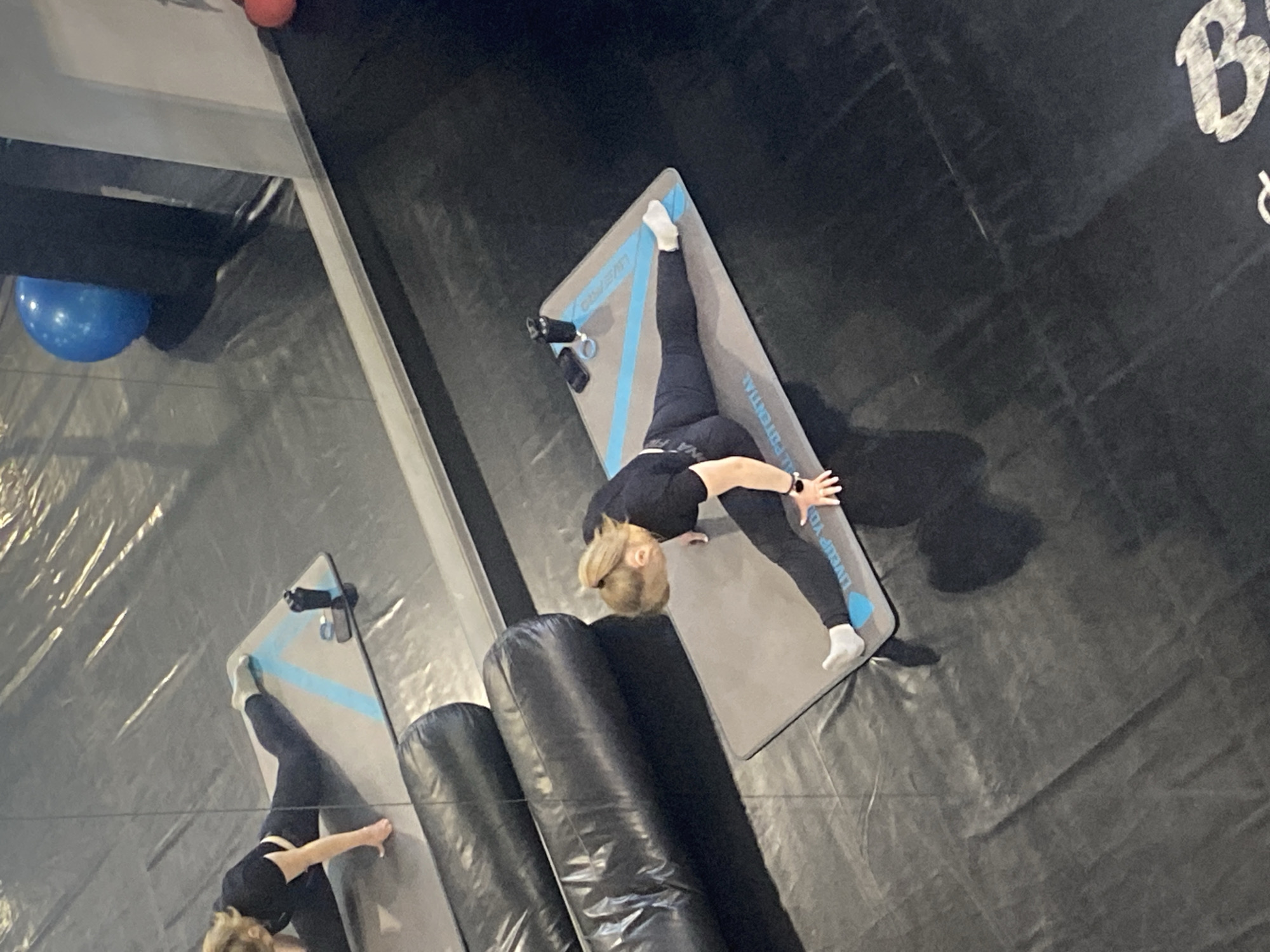A woman with blonde hair, dressed in black athletic wear and white socks, is stretching on a light gray yoga mat with a blue stripe and writing on it in what appears to be an indoor trampoline park. The mat is located on a dark trampoline surface with safety padding on one side. A mirror reflects the woman and part of the scene. A partially visible sign with the letter "B" is in the upper right. The image was not taken in Yekaterinburg, Russia.