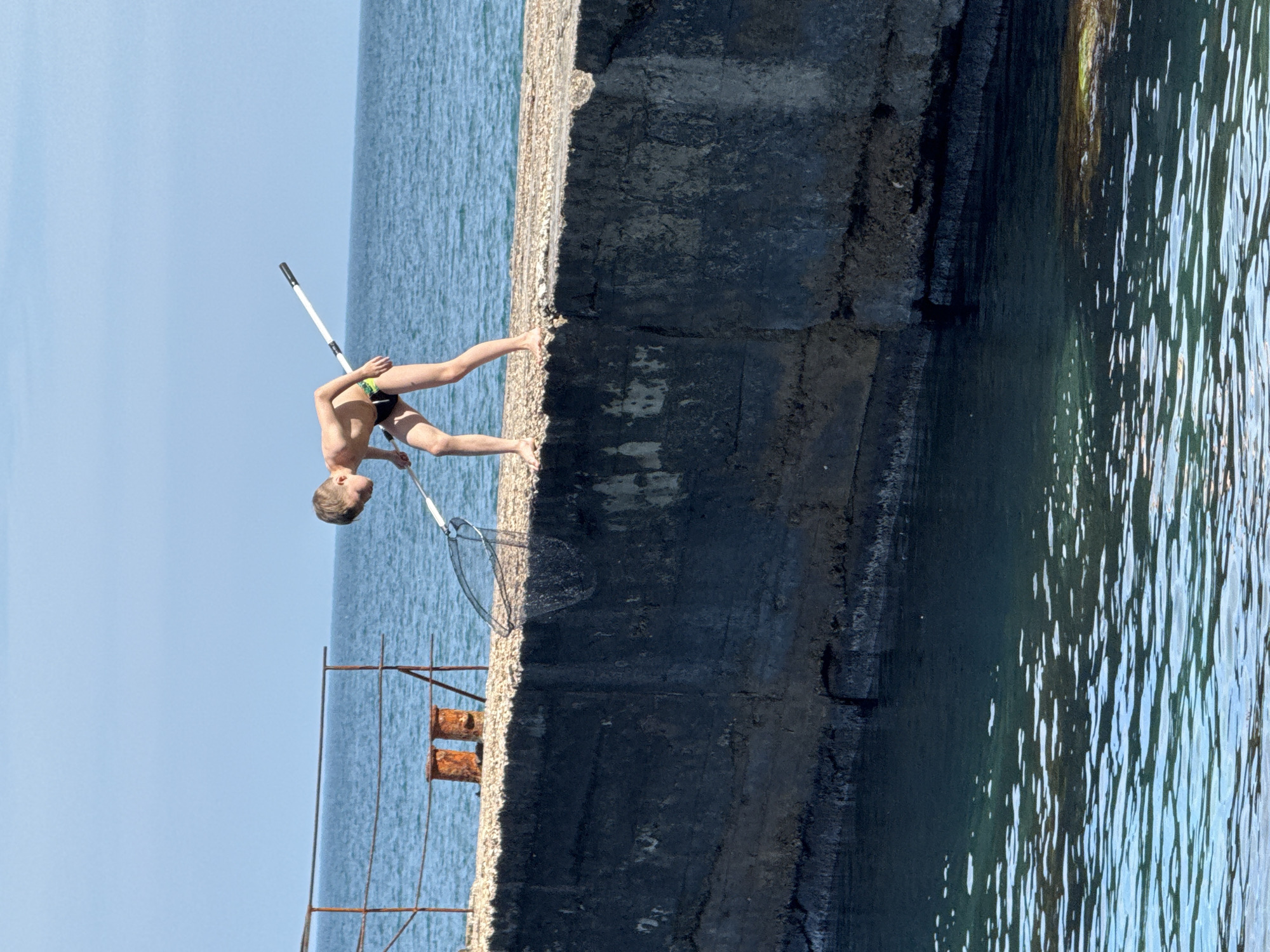 A horizontal shot shows a young person holding a pole over a concrete barrier by the water. The concrete barrier is large and dark and it is near the water. The water is light blue. On the other side of the concrete barrier is a sandy area. There is an old rusty metal fence near the boy. The sky is light blue.