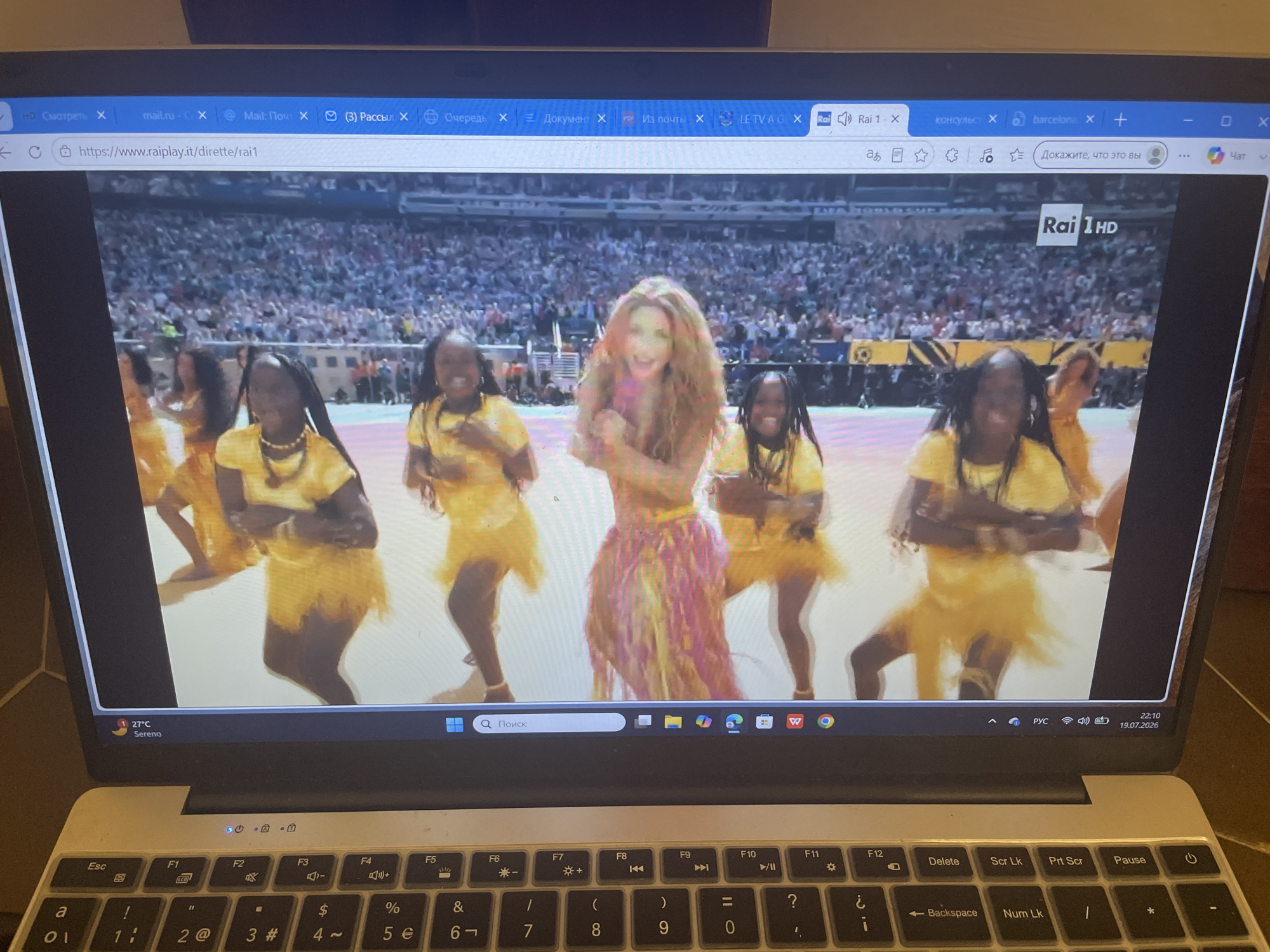 A laptop screen displays a live stream of a performance on the website `raiplay.it`. The video shows several young women in yellow outfits dancing on a stage, with a large crowd visible in the background. A woman with long, blonde, wavy hair, wearing a brightly colored dress, is in the center of the frame, also dancing. The channel displayed is "Rai 1 HD". The laptop's taskbar shows the time as 22:10 and the date as 19.07.2026. The operating system is Windows. There is no information about the city of Punta Ala, Italy, in the image.