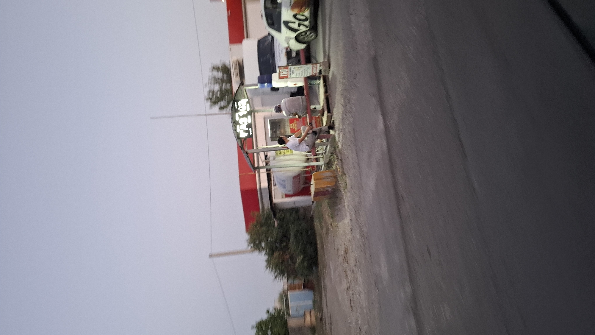 The image depicts an outdoor scene at dusk, likely near a roadside establishment in Shymkent, Kazakhstan. In the foreground, a paved road or surface is visible, with a blurred asphalt texture. In the mid-ground, a small, open-air kiosk with a red and white awning stands. Two individuals are seated on a bench in front of the kiosk. One person, wearing a light-colored t-shirt and dark pants, is looking to the right. The other, in a light-colored shirt and pants, is facing forward. Behind them, a white truck with blue containers on top is parked. To the right of the truck, a white car with a "GO" logo and what appears to be a burger graphic is visible, suggesting it might be a food delivery or fast-food service. In the background, there are trees, some blue and white structures, and what could be fuel tanks. The sky is a pale, dusky blue, indicating the late hour. The overall scene suggests a commercial area, possibly a rest stop or a food vendor location on the outskirts of the city.