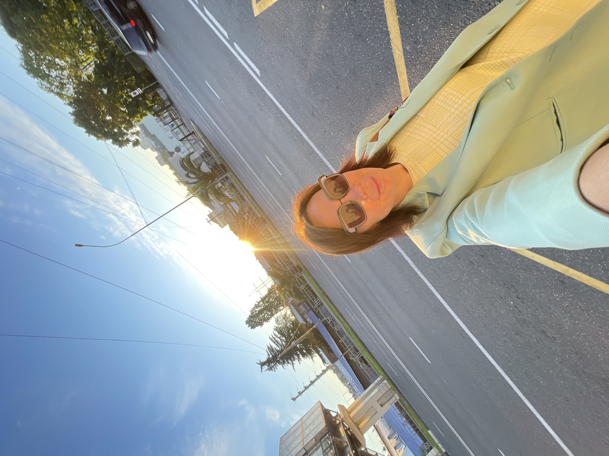 This media file presents a diagonal selfie of a woman taken outdoors on a street in Sochi, Russia, during the golden hour.
The main subject is a woman, captured from the chest up, wearing dark sunglasses that obscure her eyes. Her medium-length brown hair is backlit by the sun, creating a bright halo effect. She is dressed in a light green blazer or jacket worn over a yellow top featuring a subtle, possibly houndstooth, pattern. Her facial expression appears neutral or slightly smiling, partially obscured by the sunglasses and the intense sunlight.
The setting is an urban street or thoroughfare. The background features a paved road with distinct white lane markings and a yellow line along its edge. A dark, blurred car is visible in the distance on the road, suggesting movement. The roadside is lined with lush green trees, including prominent palm trees, alongside various urban buildings, one of which has a reflective or glass facade. Utility poles and overhead wires crisscross the clear blue sky, which has some scattered clouds. A street light fixture can also be seen against the sky.
The strong, warm sunlight, indicative of either late afternoon or early morning (golden hour), causes significant lens flare and bright glare, particularly on the left side of the image and behind the woman's head. The overall weather appears bright and sunny. There is no discernible text visible in the image.