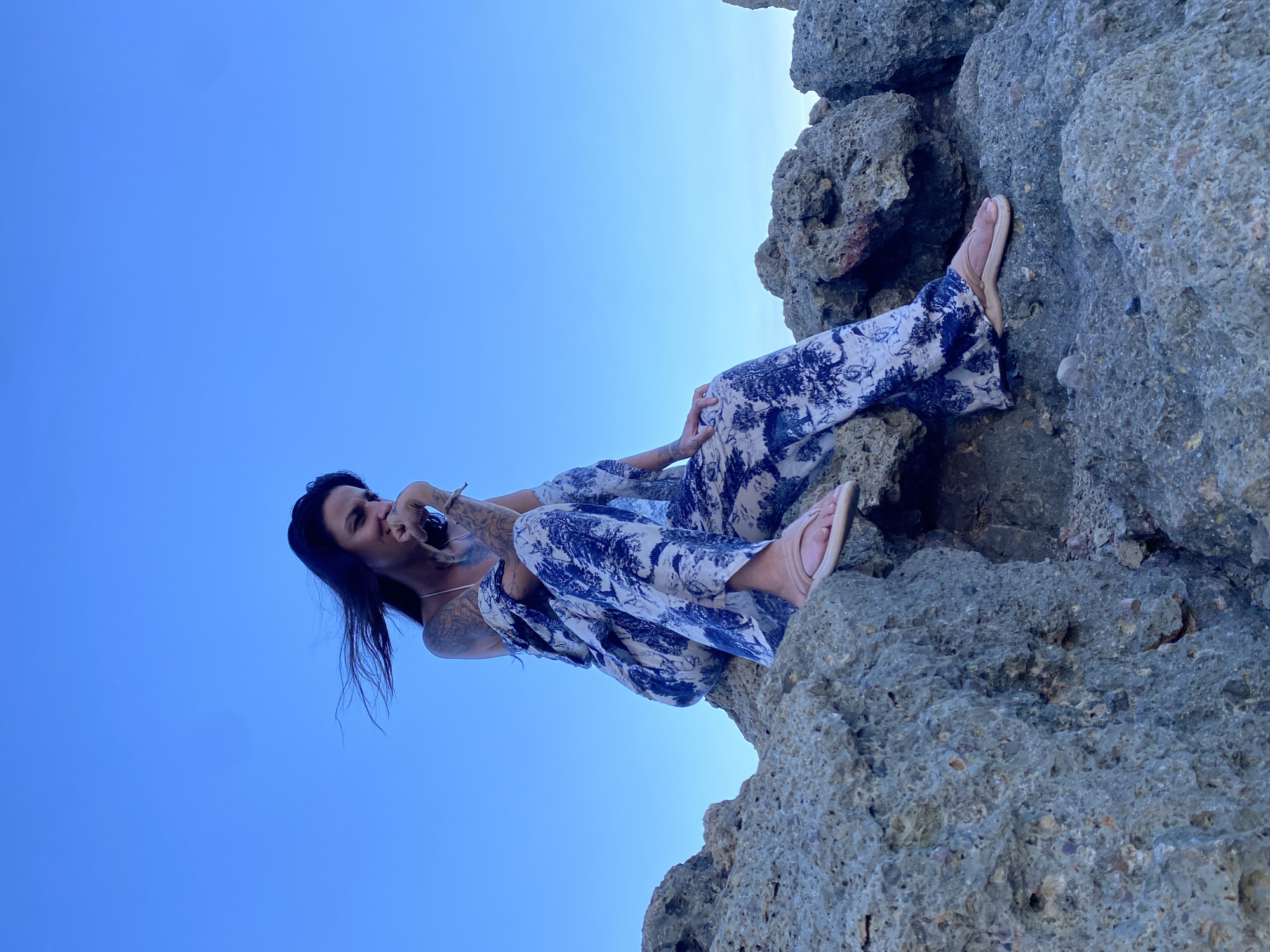 A woman with dark hair is sitting on a rocky outcrop with a clear blue sky in the background. She is wearing a patterned jumpsuit and flip-flops. Her hand is resting on her chin, and she appears to be looking down. The rocks are rough and uneven. The scene is likely located on the coast of Mui Ne, Vietnam, known for its stunning natural rock formations and beaches.