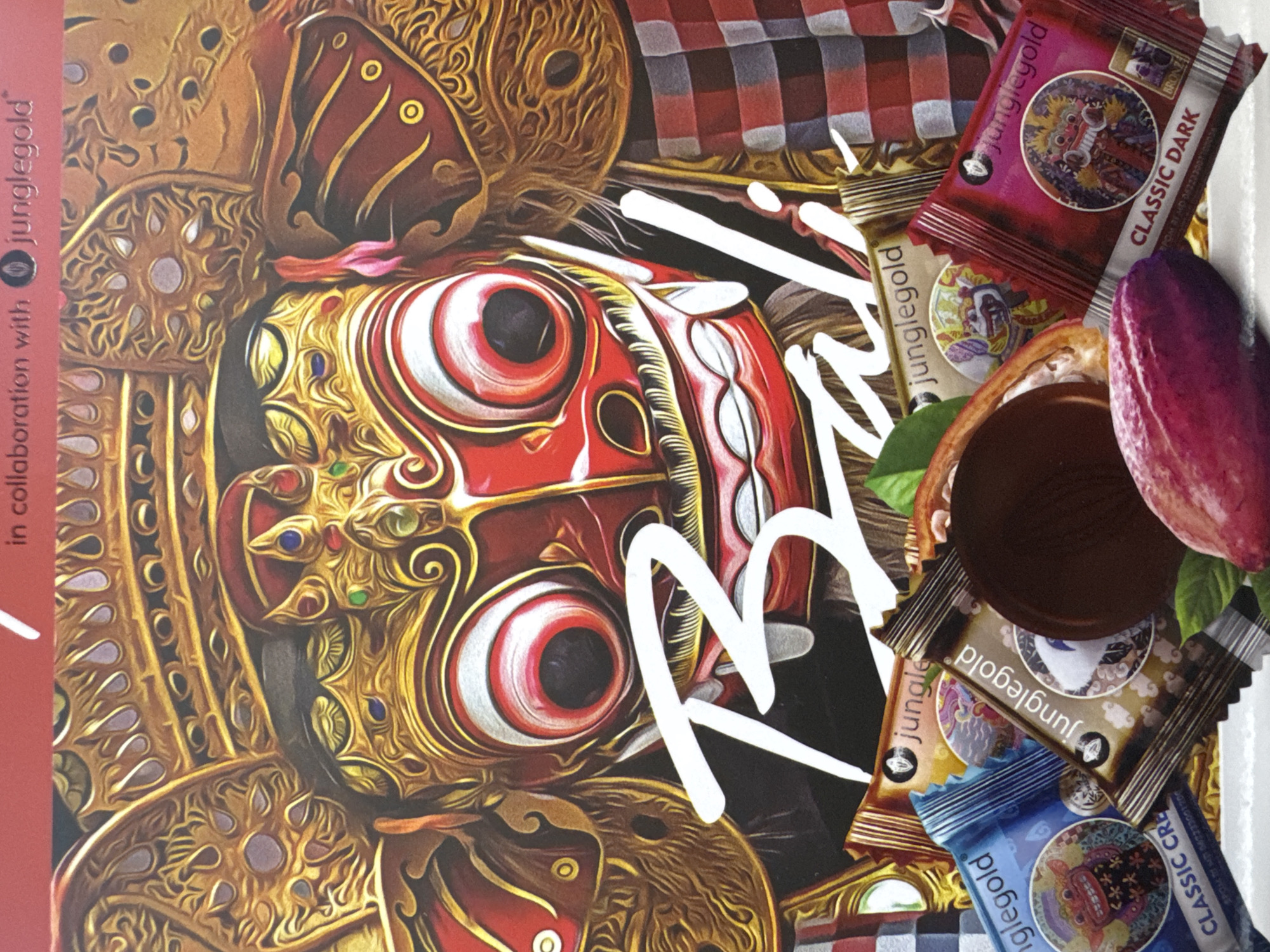 The image shows a close-up of a product featuring a Balinese Barong mask, which is characteristic of the Kuta Selatan region in Bali, Indonesia. The mask is intricately designed with gold, red, and white colors. The text "Bali" is visible in white, stylized lettering across the image. Additionally, there are several wrapped "Junglegold" chocolate bars, a chocolate candy, and a cocoa pod on the right side of the image. On the left is the text "in collaboration with Junglegold". The background consists of a red and white checkered pattern.