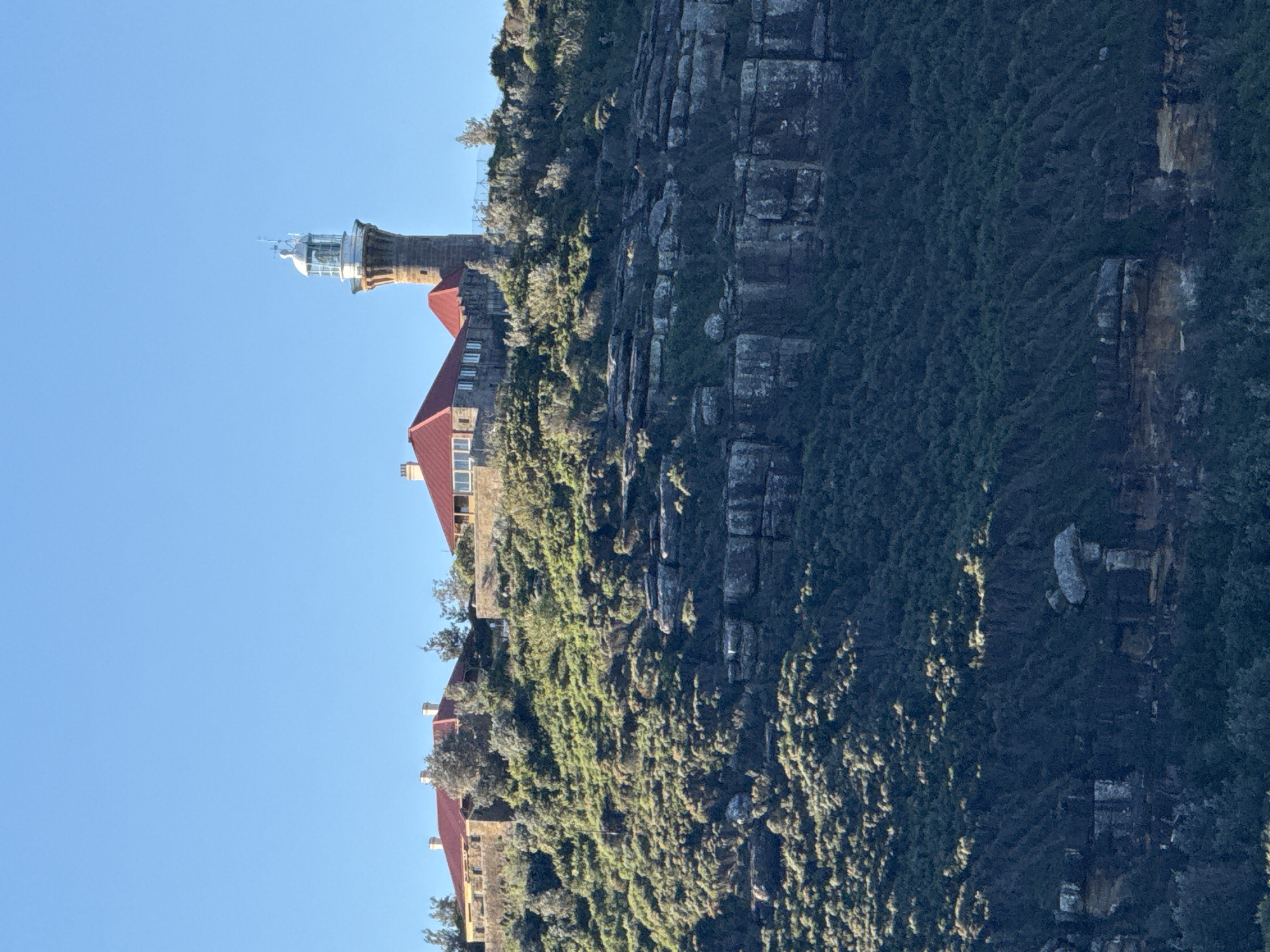This image captures Barrenjoey Lighthouse, a prominent landmark in Palm Beach, Australia. The lighthouse, constructed from stone, stands tall with a glass lantern room at its apex. It is surrounded by several stone buildings with red tiled roofs, nestled on a lush green hillside overlooking the ocean. The landscape is characterized by rocky outcrops and dense vegetation. The sky is clear and blue, indicating fair weather. The scene is devoid of any people or animals, presenting a serene and architectural view of the location.