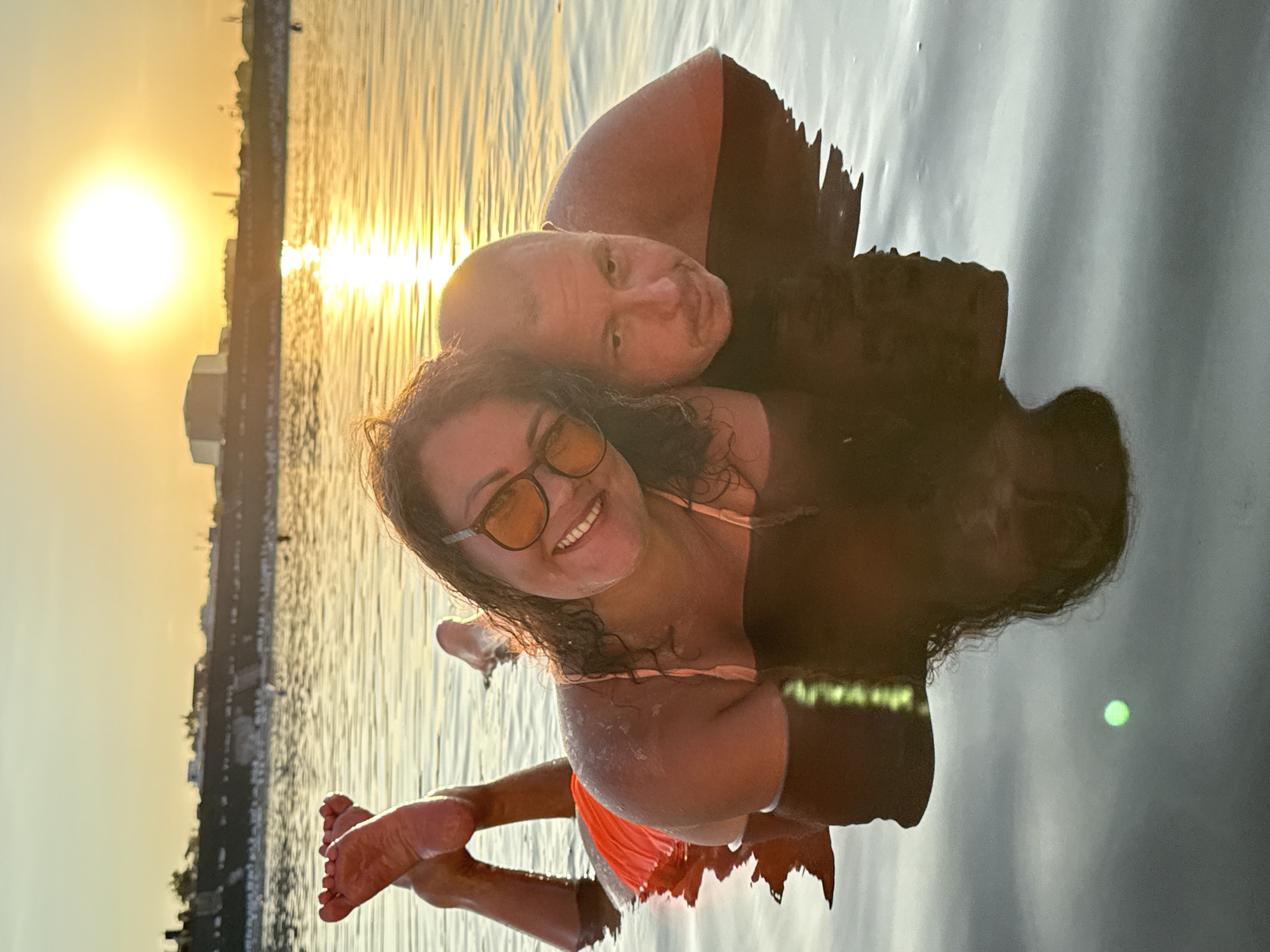 The image captures a couple relaxing in a body of water, likely at the Sol-Iletsk resort in Russia, during sunset. The woman is in the foreground, lying on her stomach with her legs crossed and lifted out of the water, facing the camera with a smile. She is wearing sunglasses and an orange swimsuit bottom. Beside her, a man rests his head on her back, looking towards the camera with a neutral expression. The water reflects the warm hues of the setting sun, which is visible in the background, casting a golden glow over the scene. The distant shoreline features buildings and a barrier, suggesting an urban or developed recreational area. The overall atmosphere is tranquil and intimate.