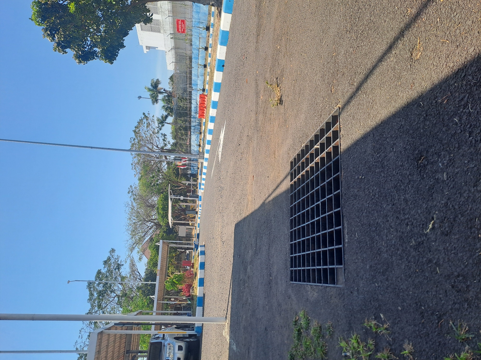 This image shows an outdoor scene in Segoro Tambak, Indonesia. In the foreground, a metal grate is set into the asphalt of a paved area, likely a road or parking lot. To the left of the grate, a small patch of green grass is visible. In the middle ground, there are several covered parking structures with white roofs supported by metal poles. A silver SUV is partially visible under one of these structures. The paved area is bordered by a low wall painted with blue and white stripes. Beyond the wall, a fence encloses a white building with a flat roof and barred windows. Trees and palm trees are scattered throughout the scene. The sky is clear and blue, suggesting a sunny day. A long shadow from an unseen object stretches across the paved surface. There are no people visible in the image.