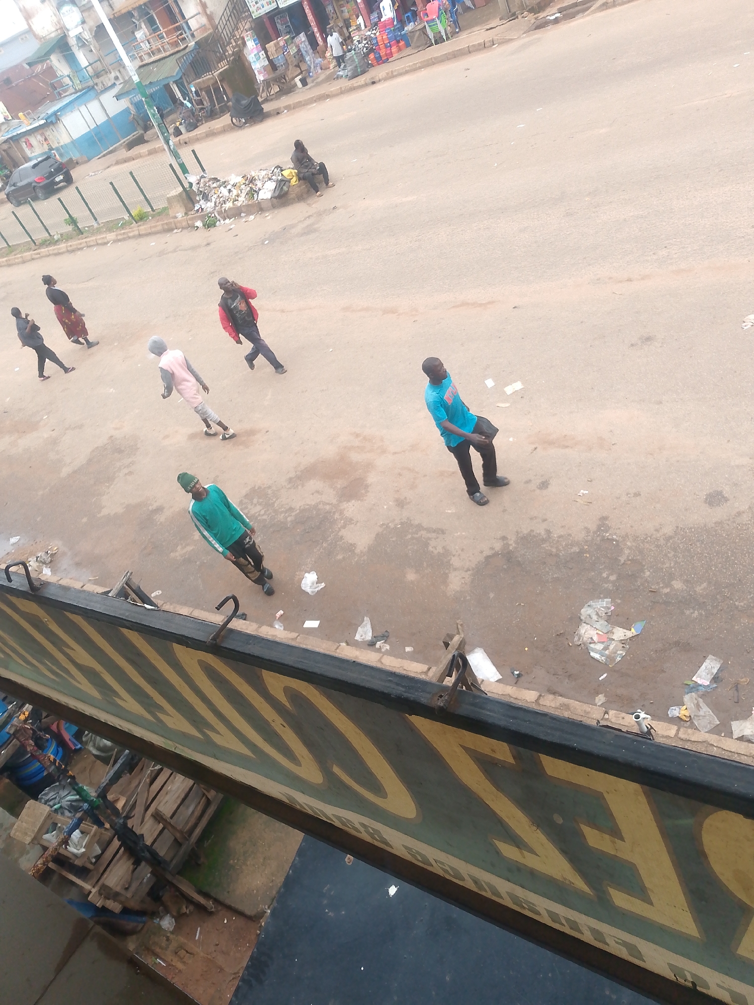 This aerial photograph, taken in Bukuru, Nigeria, captures a street scene with several individuals walking and standing on a paved road and adjacent dirt areas. In the foreground, a dark sign with yellow lettering is visible. The street is lined with low-rise buildings, some appearing to be shops or residential structures, with awnings and visible goods outside. A black car is parked on the side of the road. Several people are scattered across the image; one person in a blue t-shirt and dark pants walks away from the camera, another in a green long-sleeved shirt and dark pants stands looking towards the camera, and others are seen in various colored clothing walking or standing further in the distance. Piles of trash are visible on the side of the road near a fence. The overall environment suggests a bustling urban area with some areas of litter and modest infrastructure.